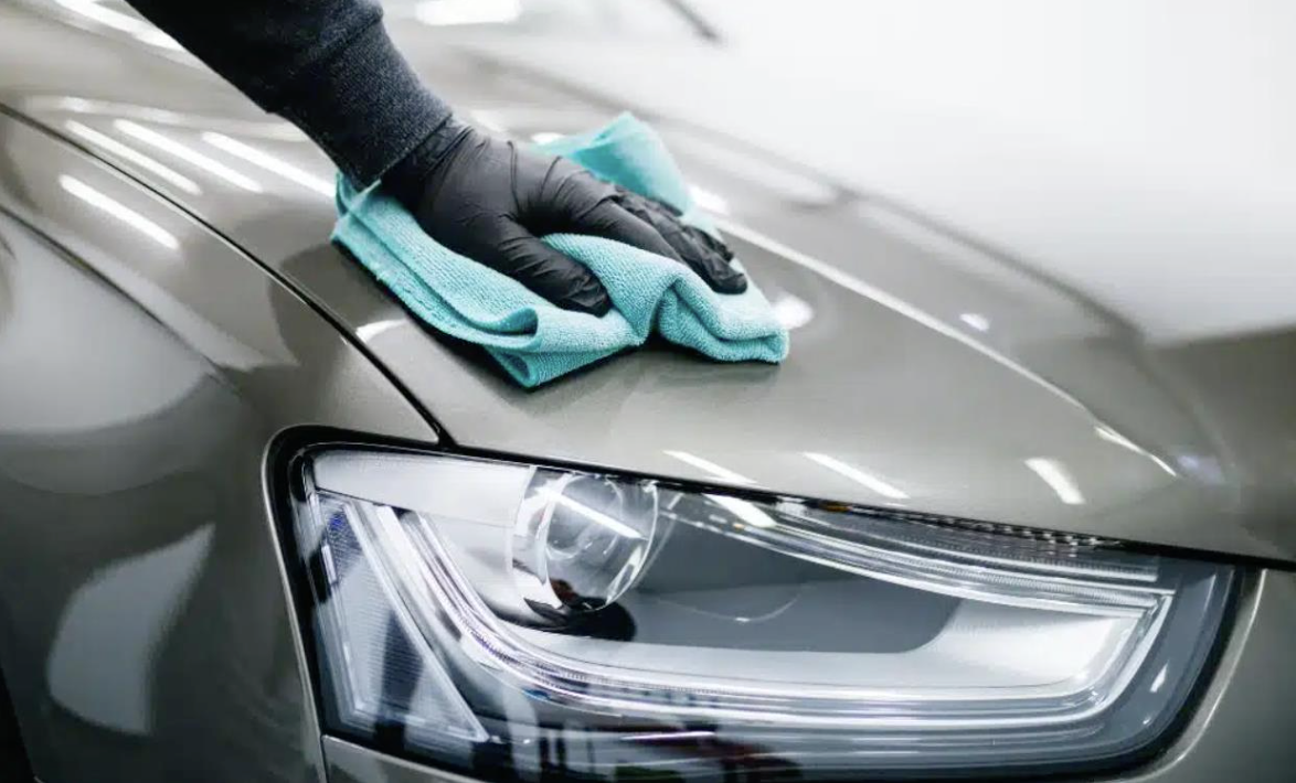 Person wearing black gloves cleaning the hood of a gray car with a blue cloth.