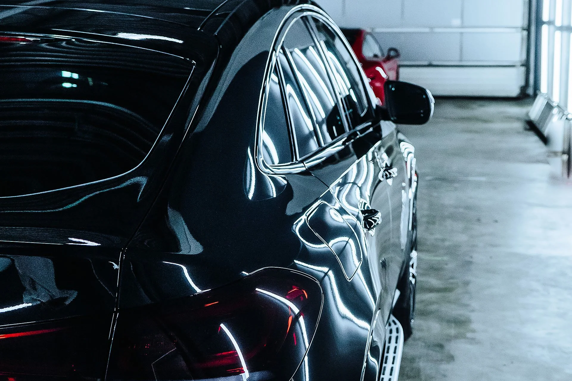 The image shows a black luxury car parked inside a garage, with a red car visible in the background.