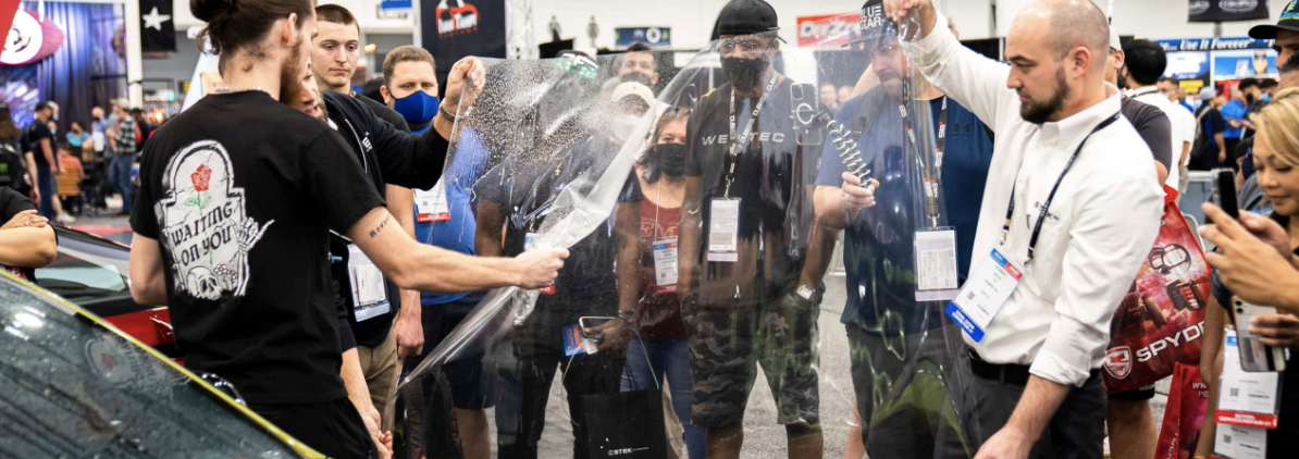 Group of people demonstrating the use of a clear protective windshield at an indoor event, with some watching and taking photos.