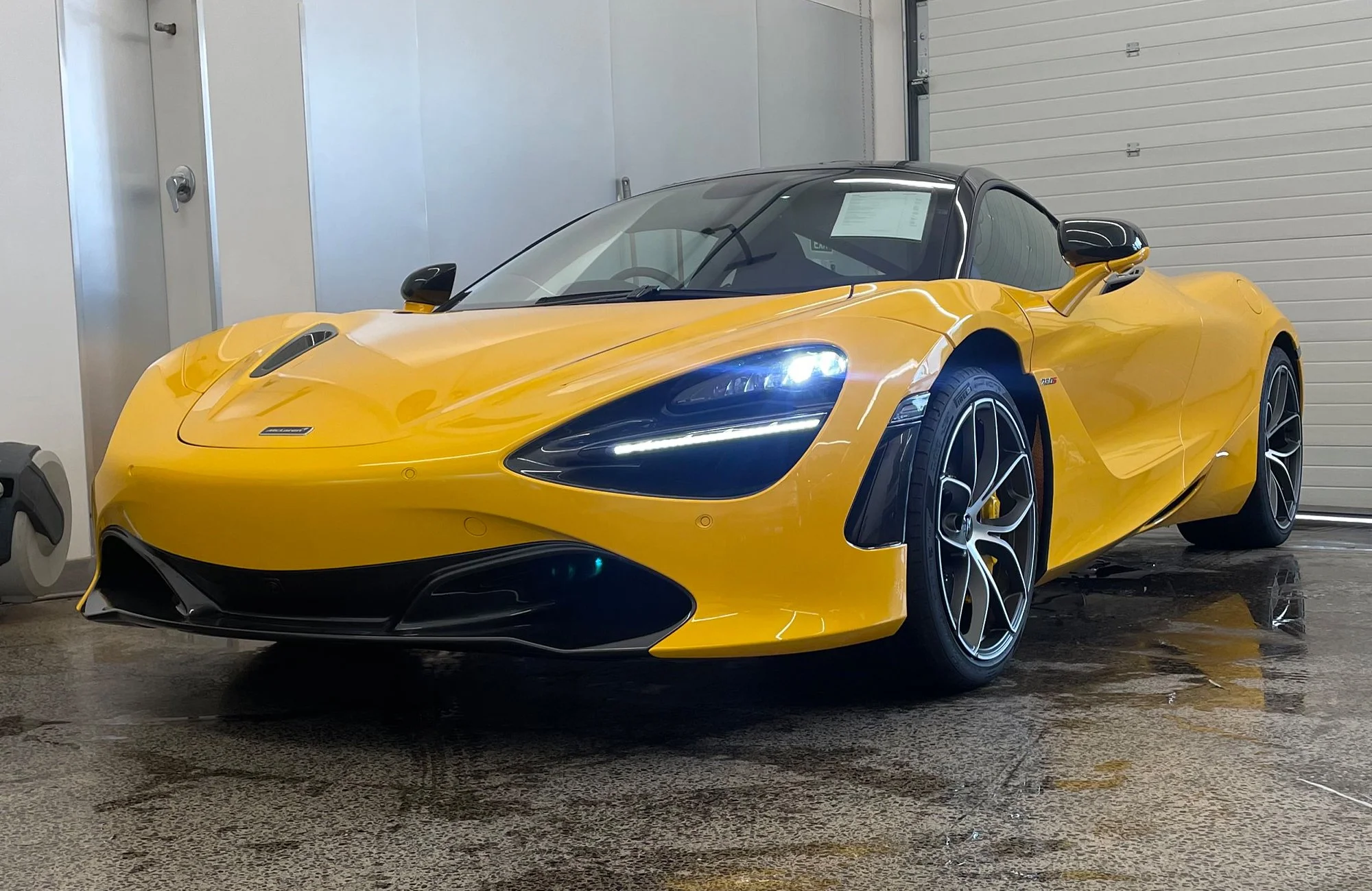 Yellow sports car with black accents parked indoors, front headlights on, and reflection on wet floor.