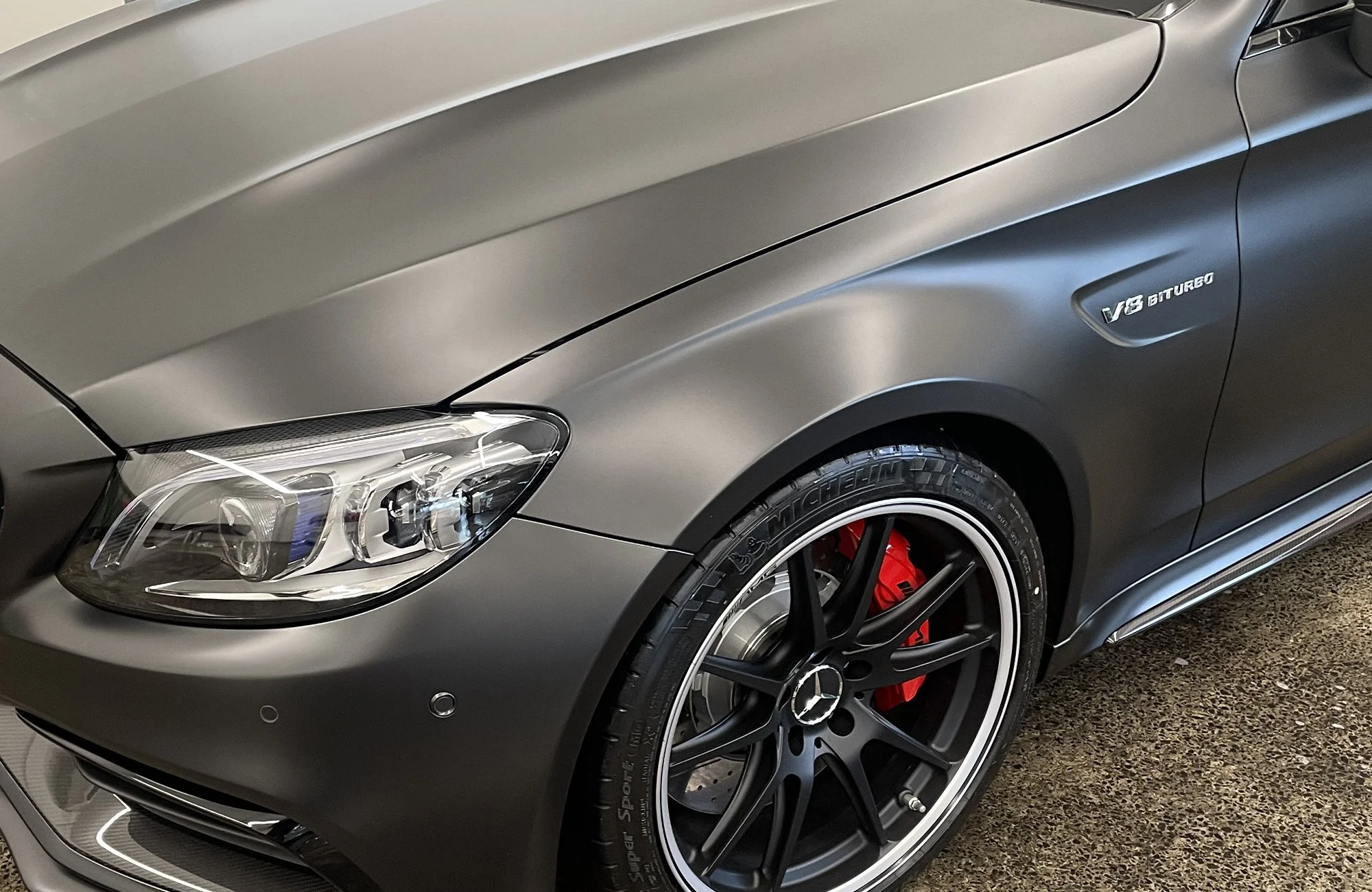 Close-up of a matte gray Mercedes-Benz car with AMG V8 bi-turbo badge, black alloy wheel with red brake caliper, and Michelin tire.