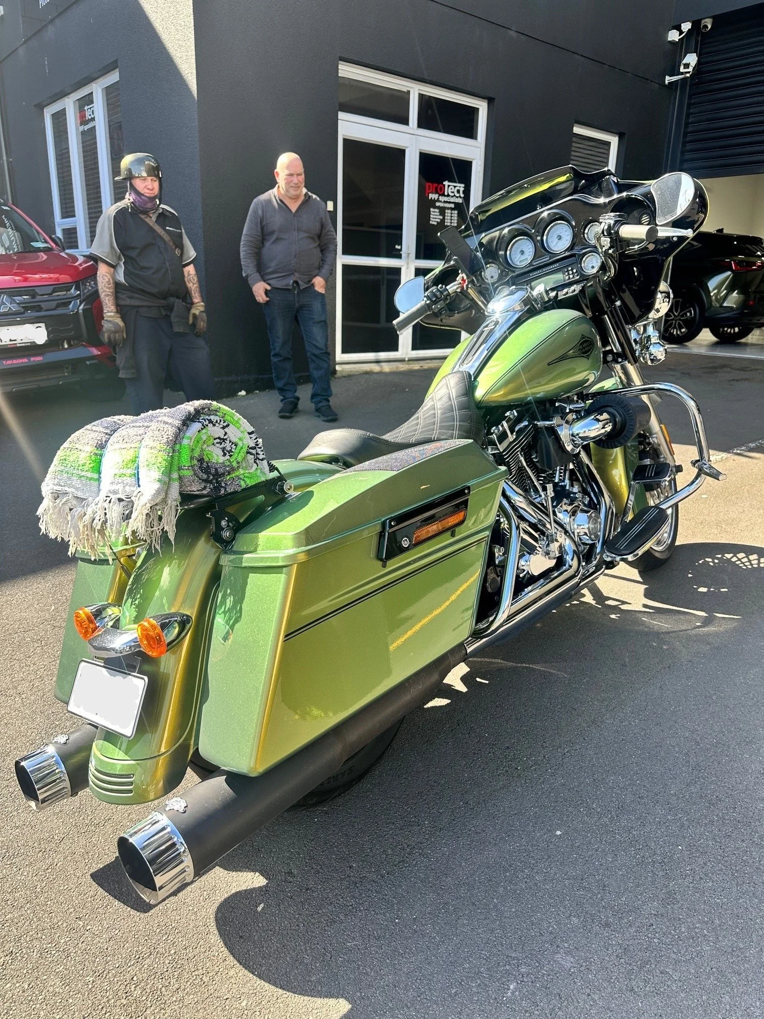 Green motorcycle with saddlebags and exhaust pipes parked outside a building, with two men standing in the background.