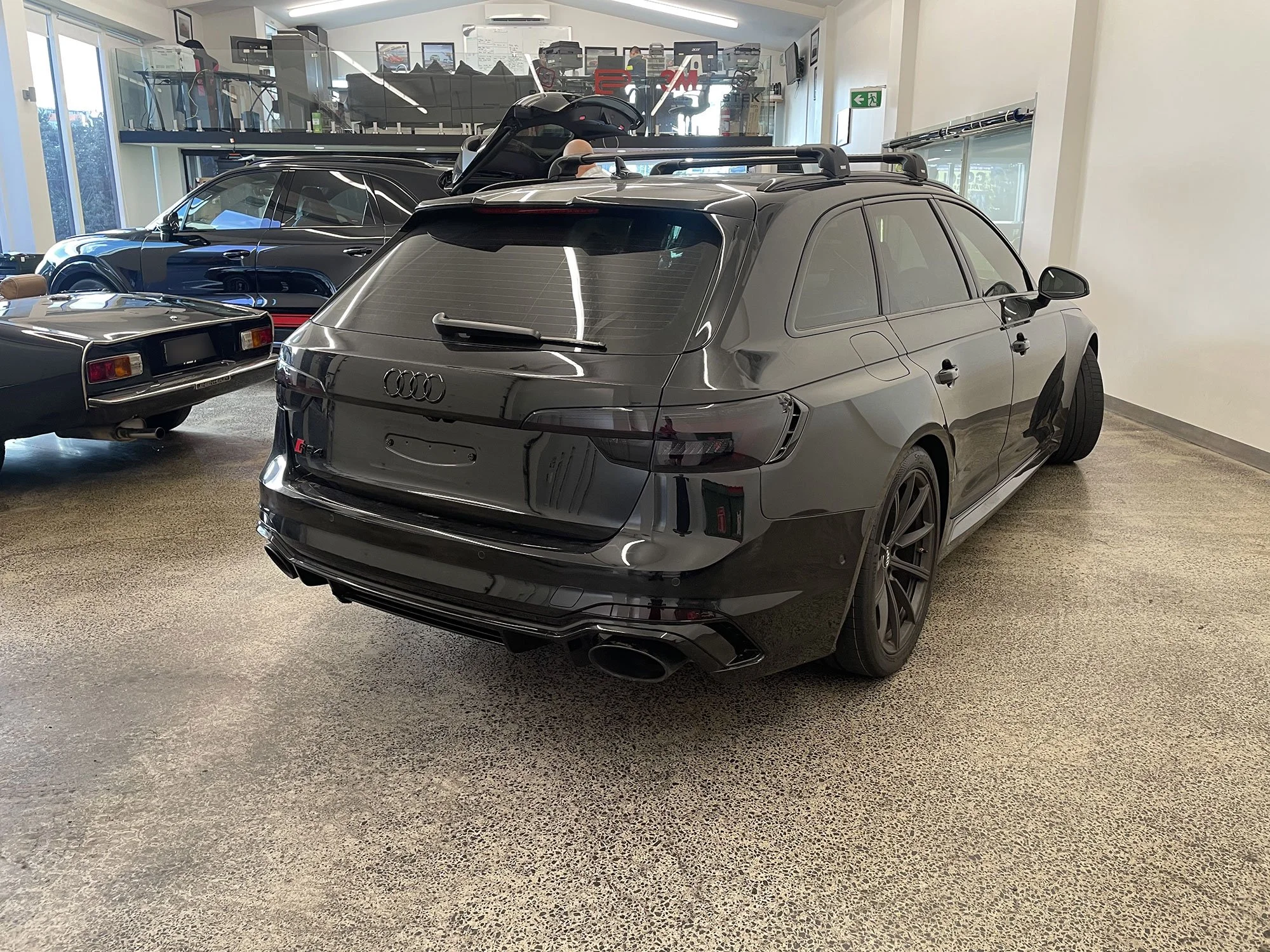 A black Audi station wagon inside a garage, surrounded by other classic and modern cars, with a bicycle mounted on the wall and indoor showroom displays.