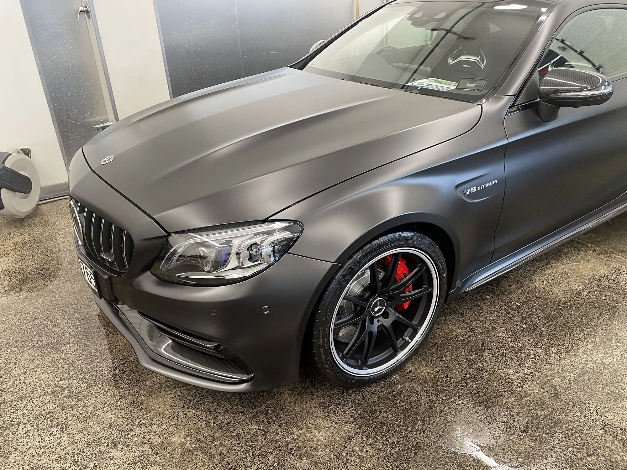 A matte gray Mercedes-Benz car parked inside a garage, featuring black wheels with red brake calipers, and a badge indicating it is a V8 Biturbo model.