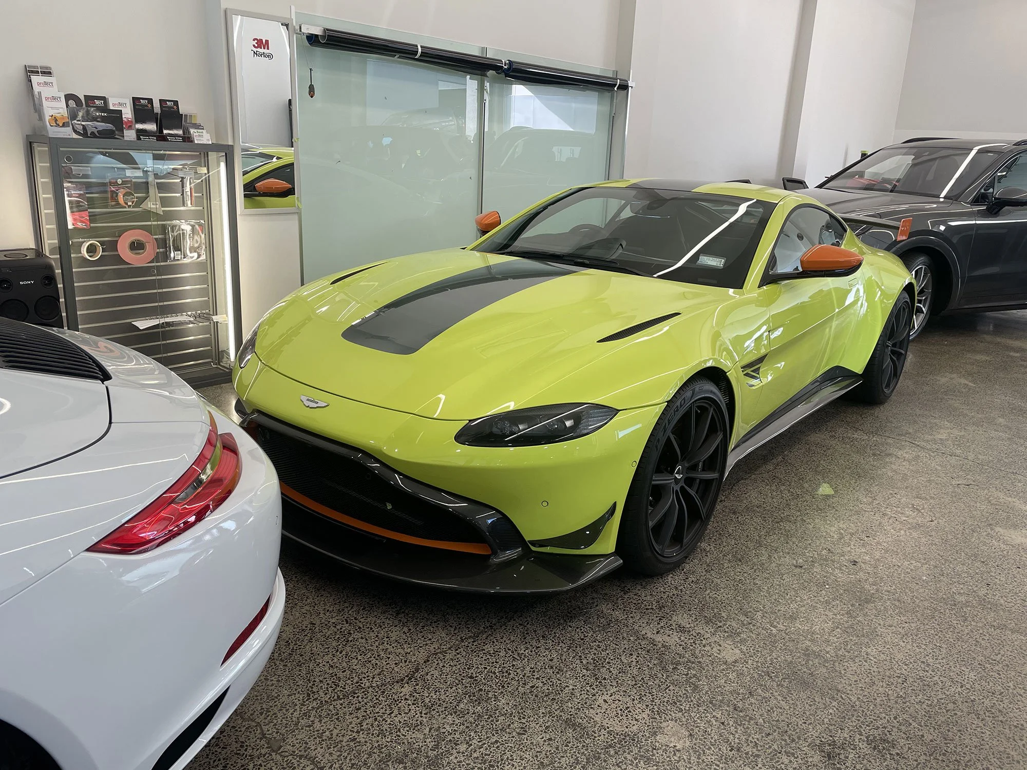 A lime green Aston Martin sports car with black details and orange side mirrors parked indoors among other cars.