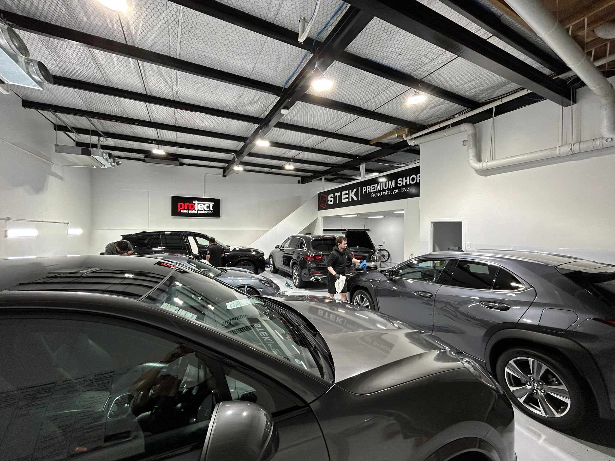 Interior of an auto detailing shop with several cars being serviced, including a focus on two technicians working on vehicles in the background. The shop has a modern look with white walls, industrial ceiling, and signage for protect, STEK, and premi