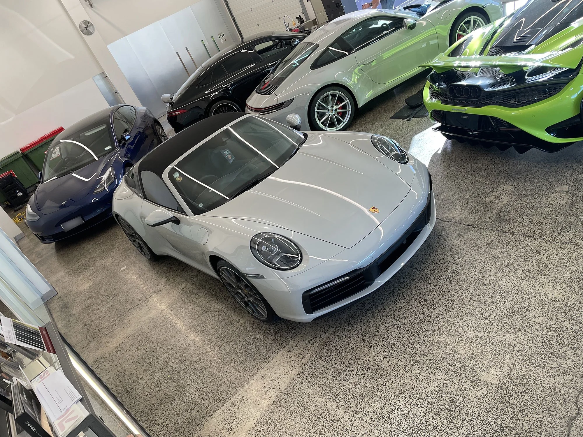 A showroom display of luxury sports cars, including a silver Porsche 911, a green Lamborghini Huracan, a black Porsche 911, a dark blue Porsche Taycan, and a white Porsche 911 Carrera convertible.