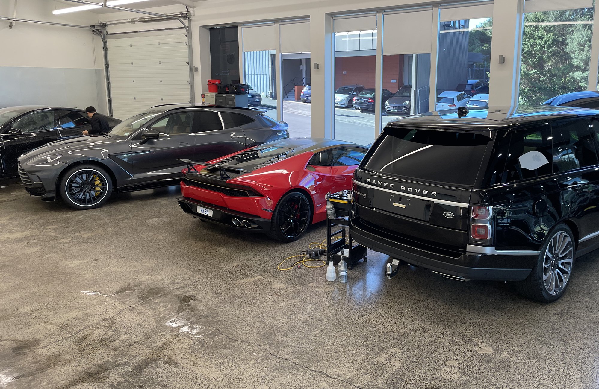 Indoor car storage with three luxury vehicles: a black Aston Martin, a red Lamborghini, and a black Range Rover, with a mechanic working on the Aston Martin.