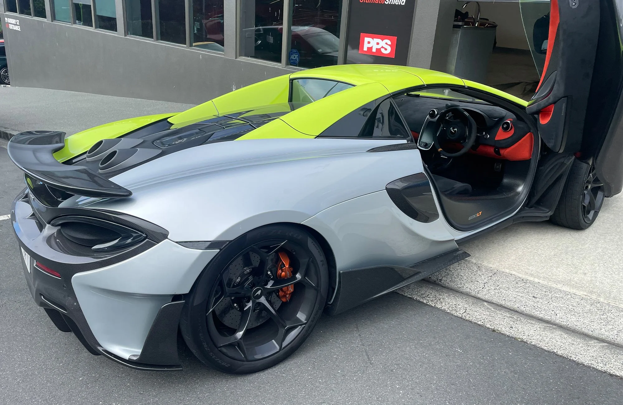 A silver and black McLaren Senna parked with the driver side door open, revealing a black and red interior, with a lime green sports car parked behind it.