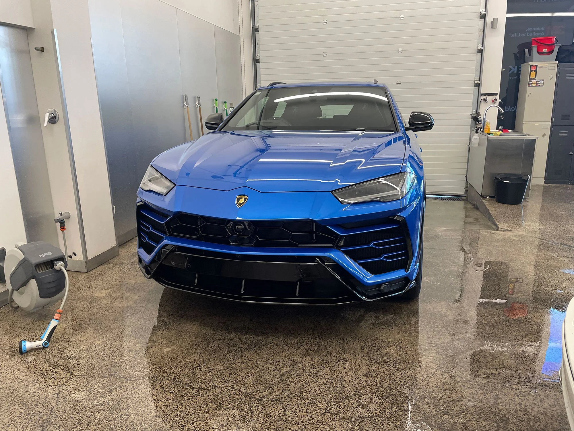 Blue Lamborghini parked inside a garage with a polished concrete floor, storage cabinets, cleaning supplies, and utility sink in the background.