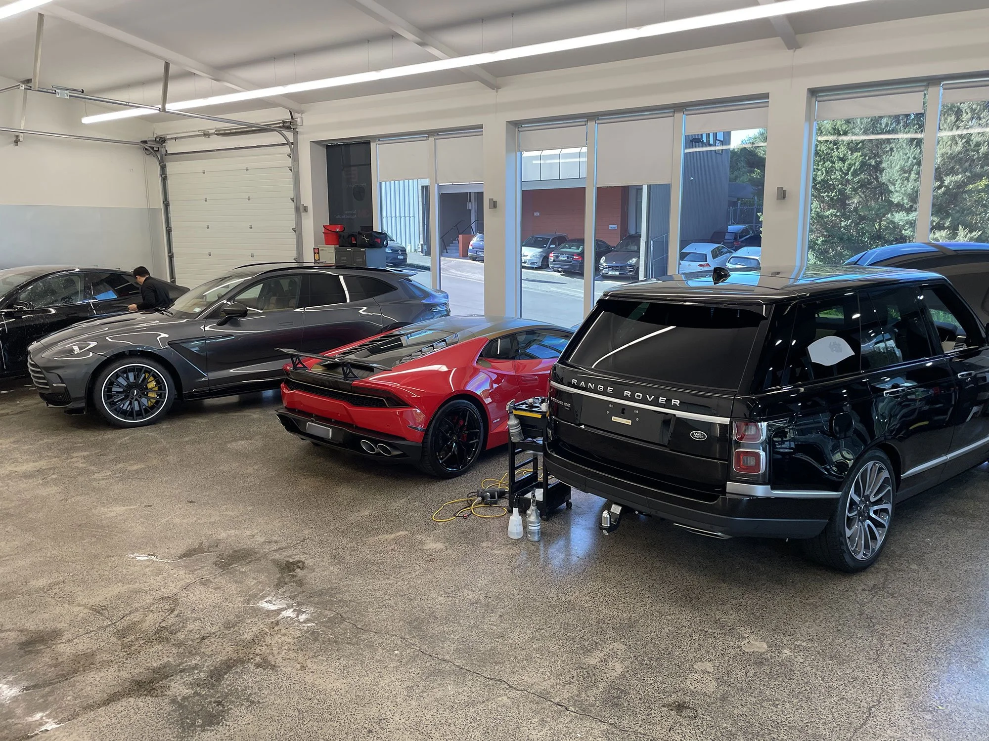 Inside a car dealership or garage, a black Range Rover, a red sports car, and a black sedan are parked. Large windows reveal an outdoor parking lot with parked cars and trees.