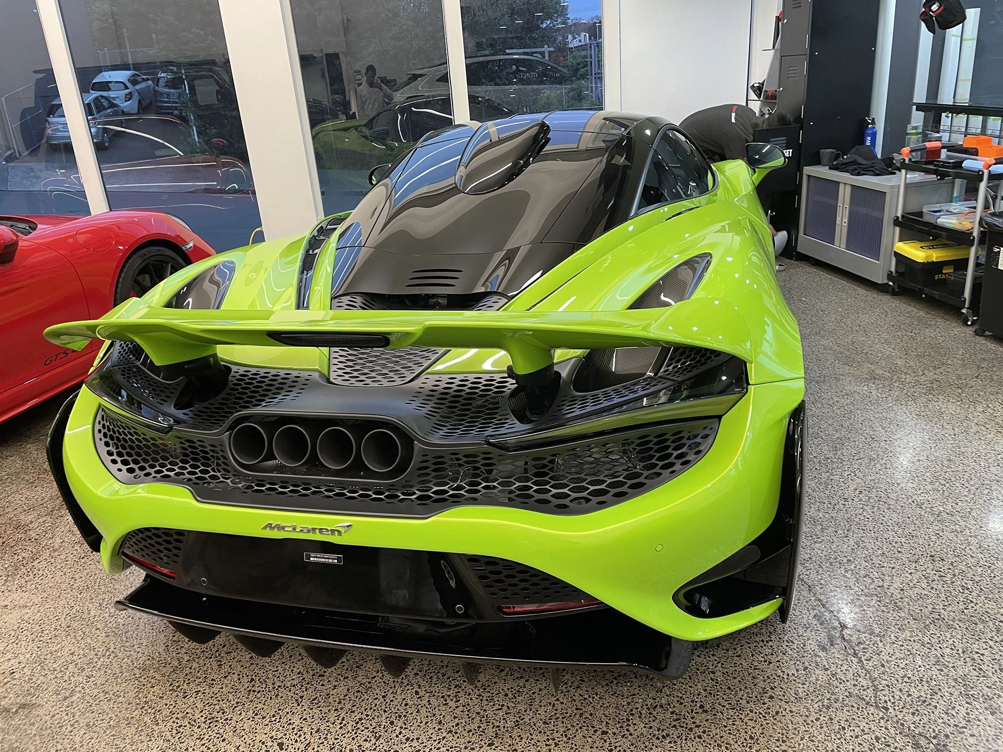 A bright green McLaren racing car inside a showroom, with a red sports car next to it and large windows in the background showing parked cars outside.
