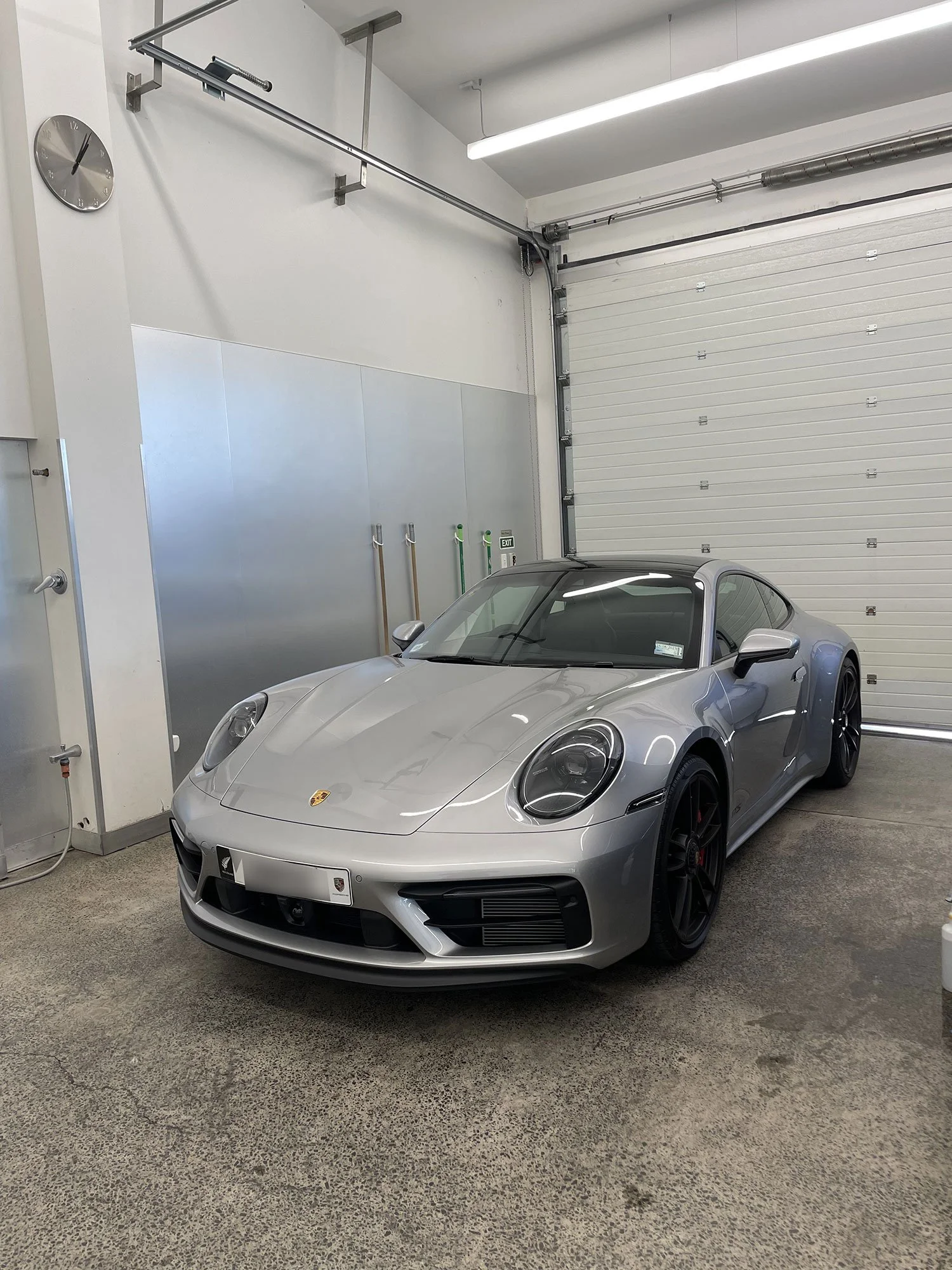 Silver sports car, a Porsche 911, parked inside a clean, industrial garage with a roll-up door and minimal decor.
