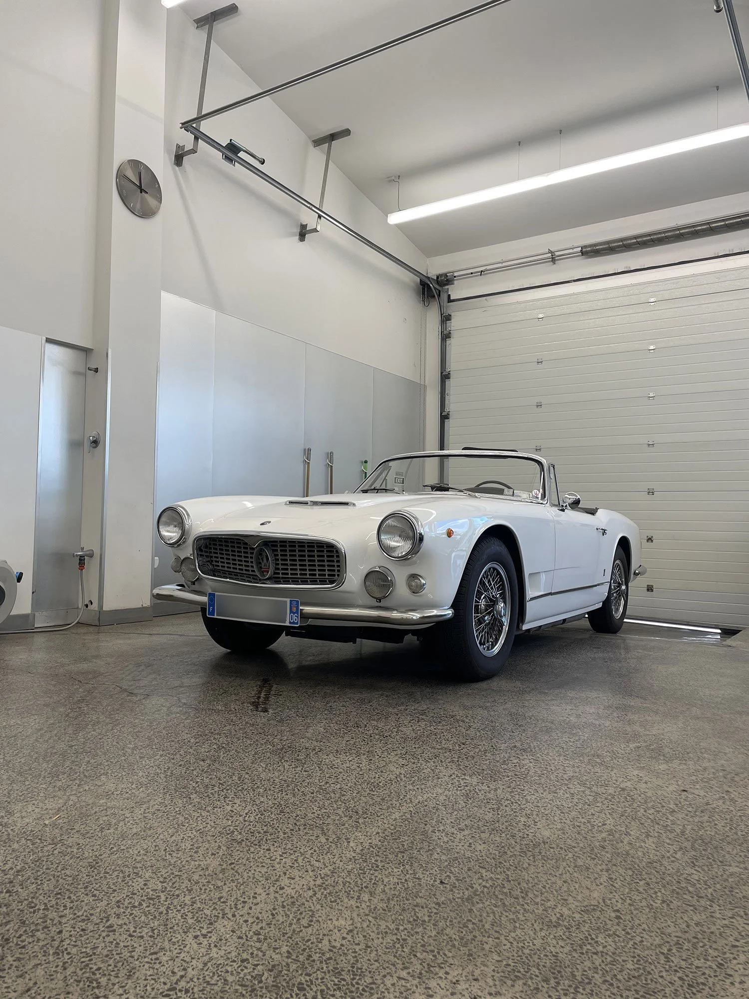 A white vintage convertible sports car parked inside a garage with a metallic roll-up door, a clock on the wall, and bright overhead lighting.