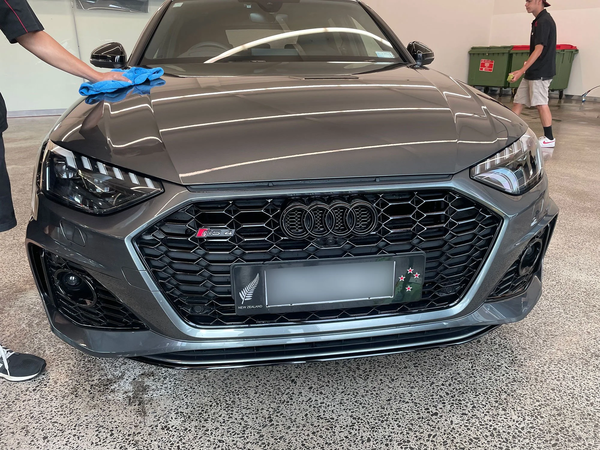 Front view of a black Audi RS4 car inside a garage, with a person cleaning the hood with a blue cloth on the left, and another person walking in the background with a green sponge and wearing a black shirt and beige shorts.