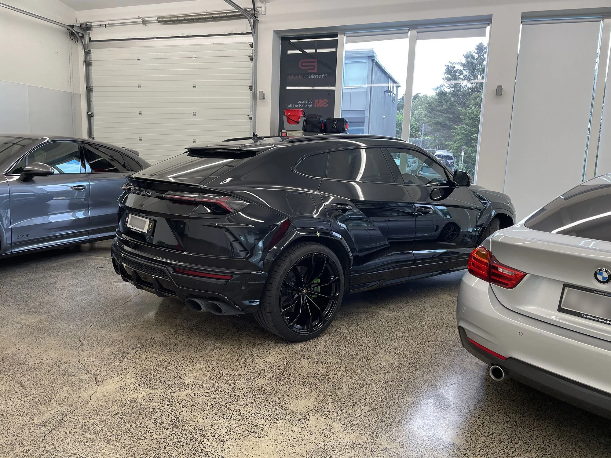 Black luxury SUV parked between a gray sports car and a white sedan inside a garage with large windows and concrete floor.