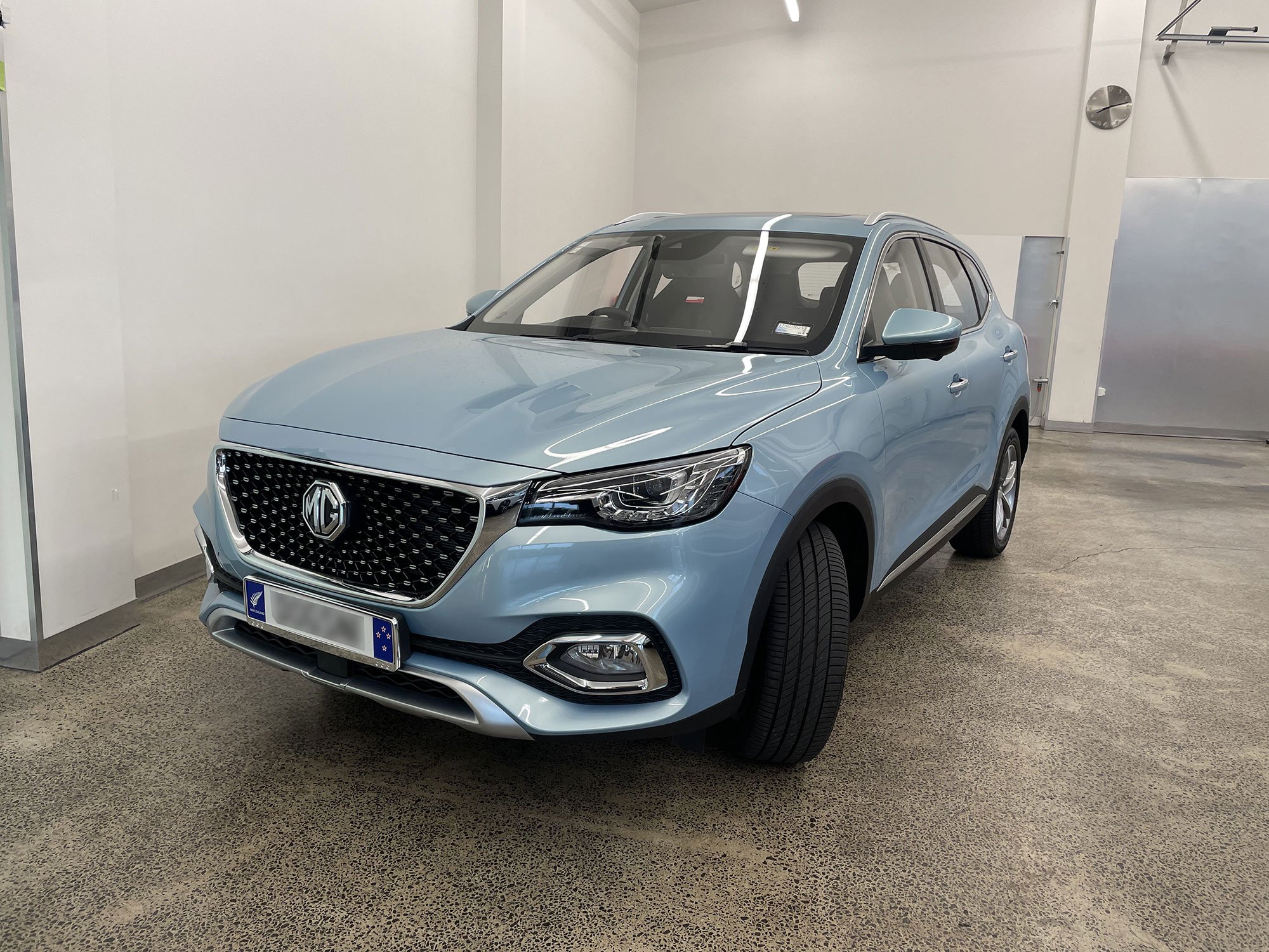 A light blue MG SUV parked indoors on a speckled brown floor, with a plain white wall and metallic door in the background.
