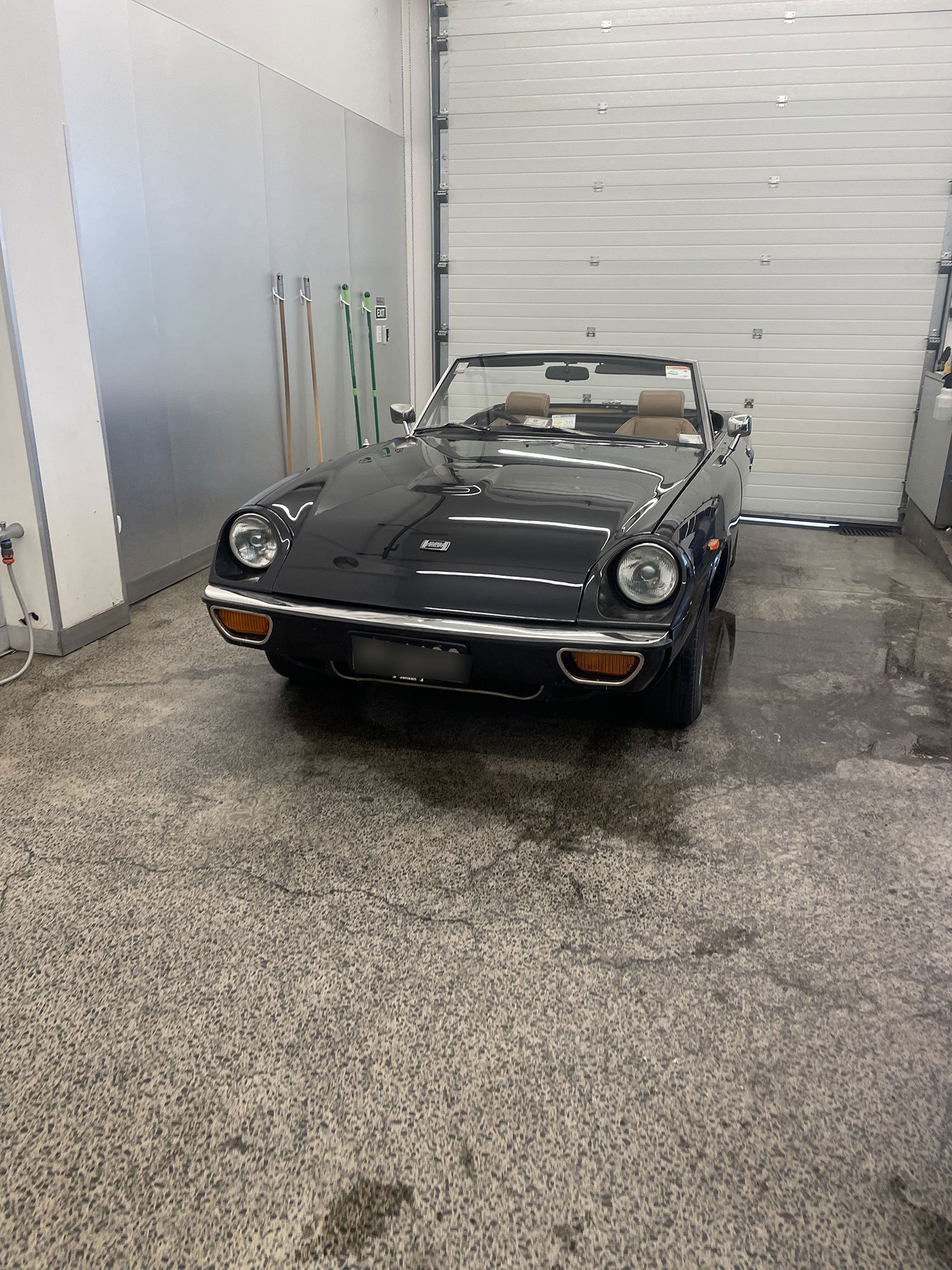 A black vintage convertible car parked inside a garage with a rolling door and cleaning tools on the wall.