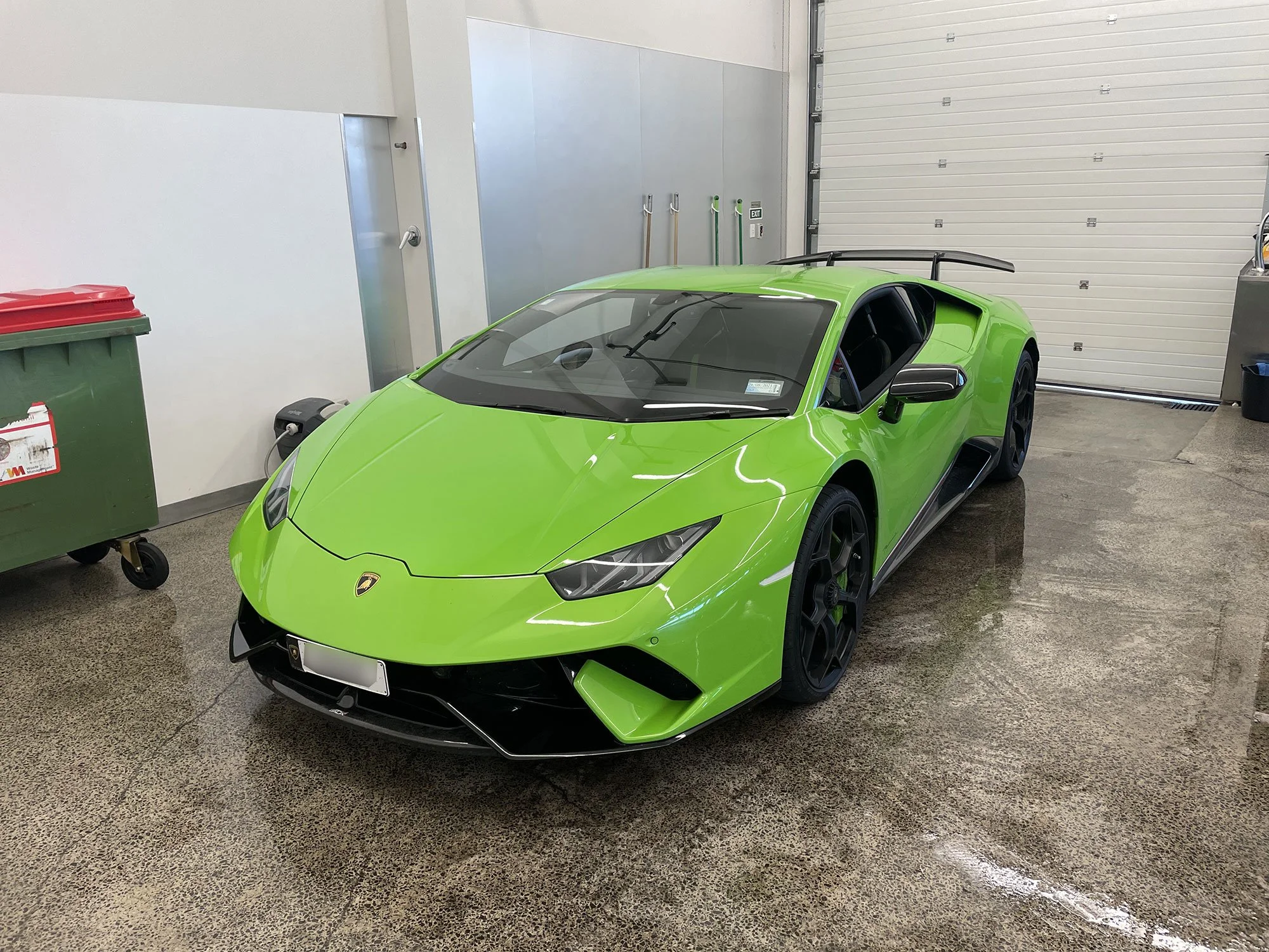 A bright green Lamborghini sports car parked inside a garage with a polished concrete floor, white walls, and a large garage door in the background.
