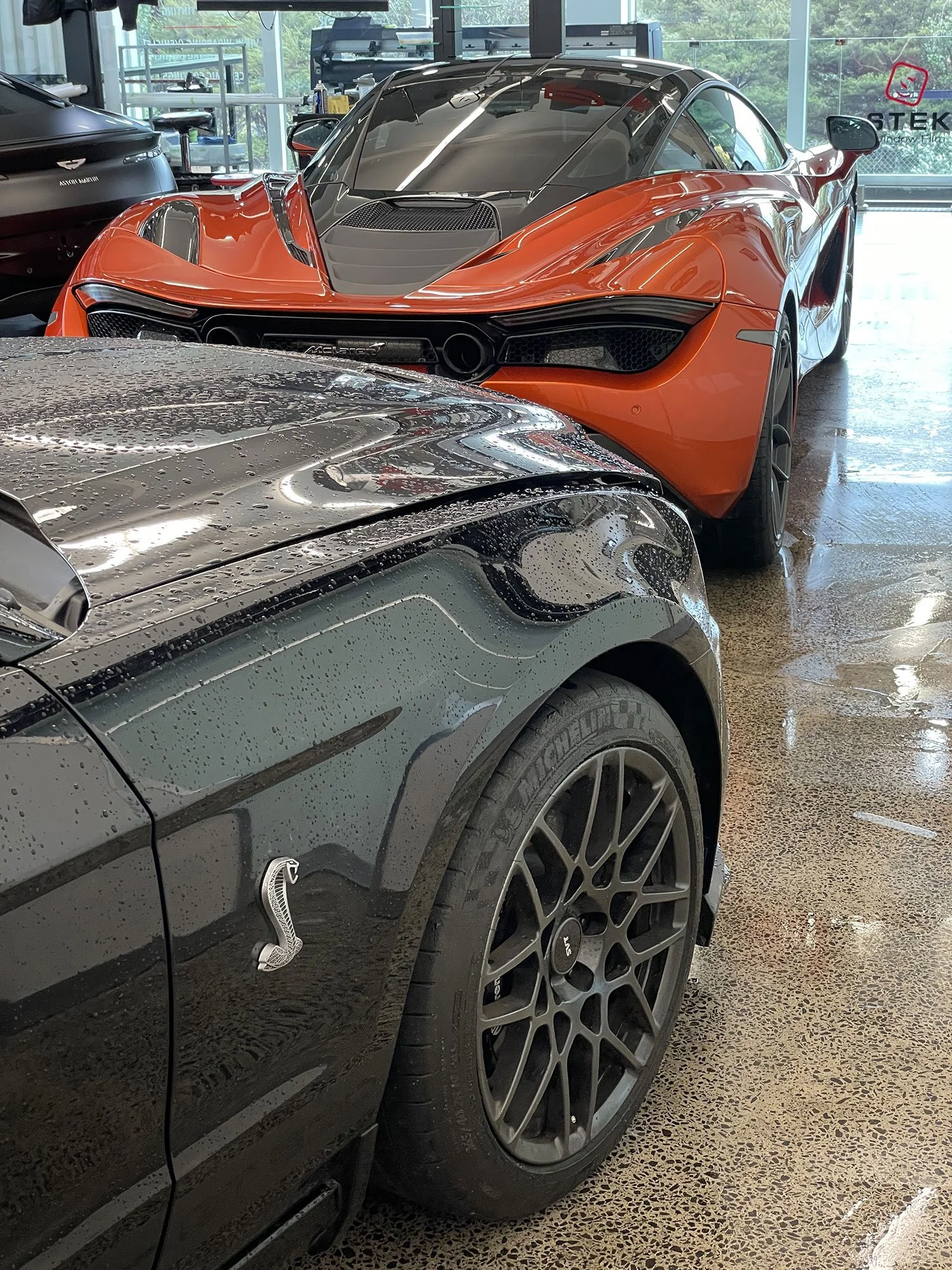 A black Ford Mustang Shelby GT350 with a Cobra emblem on the side, parked next to a red McLaren sports car inside a showroom or garage, with wet floor and water droplets on the cars.