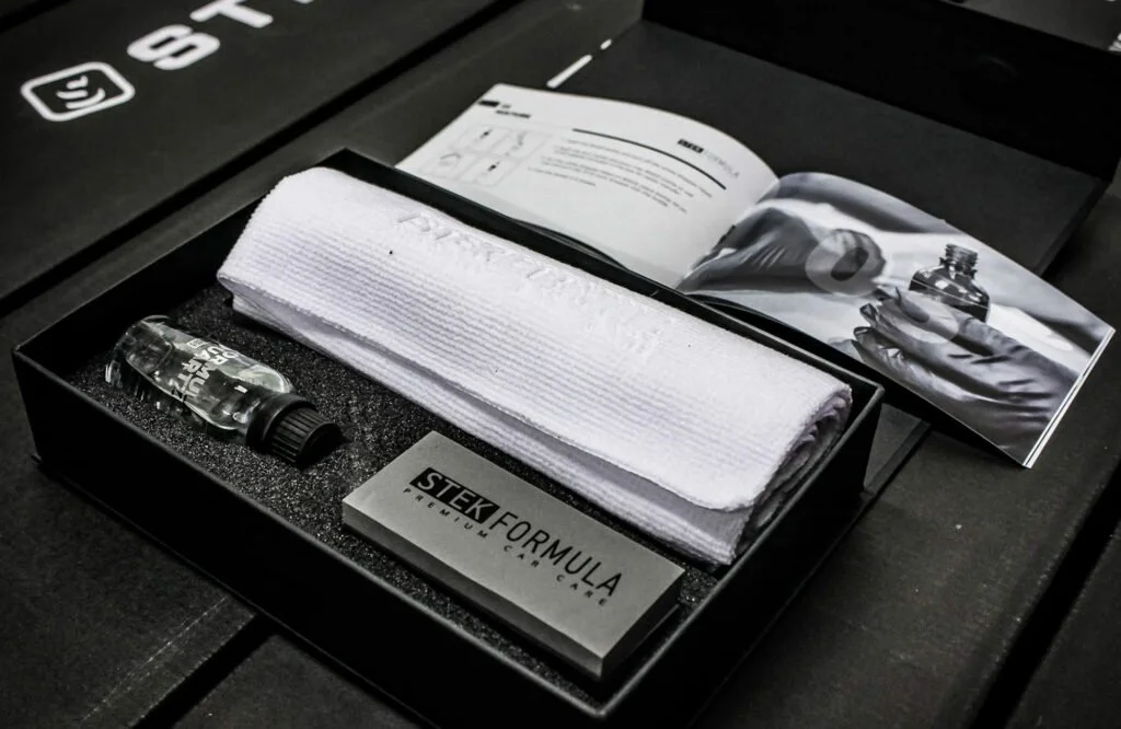 A skincare or beauty product starter kit with a small bottle, a rolled white cloth, a metallic box labeled "STEK FORMULA," and an open instruction booklet, all arranged in a black box.