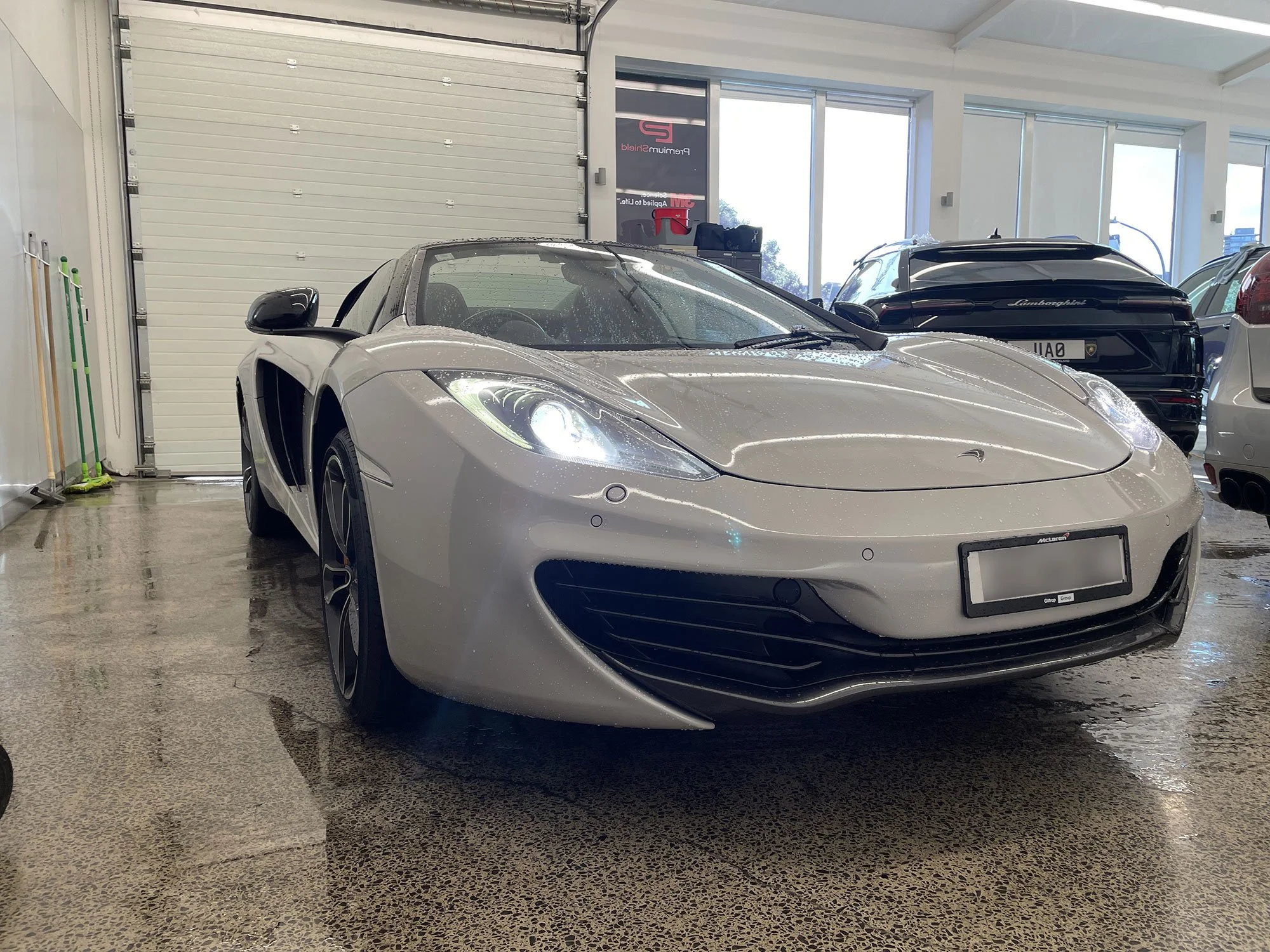 A silver McLaren sports car parked indoors with water droplets on its surface, surrounded by other high-end cars in a well-lit showroom.