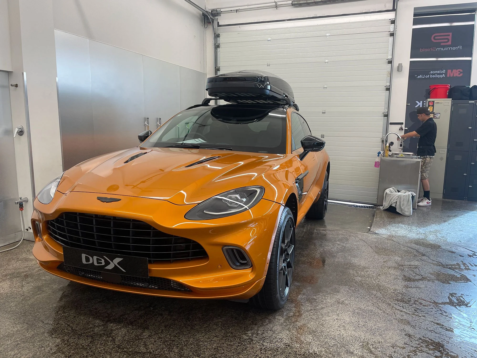 An orange sports car with a black roof box inside a garage, with a person washing hands at a sink in the background.