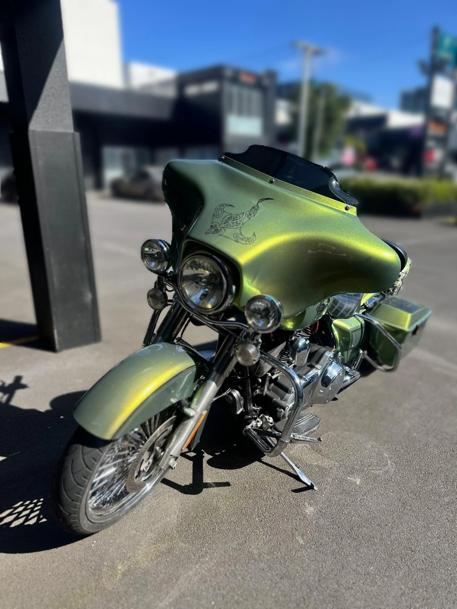Green motorcycle with a black windshield and tribal design decal on the front fairing parked on asphalt, with a blurred background of buildings and street.