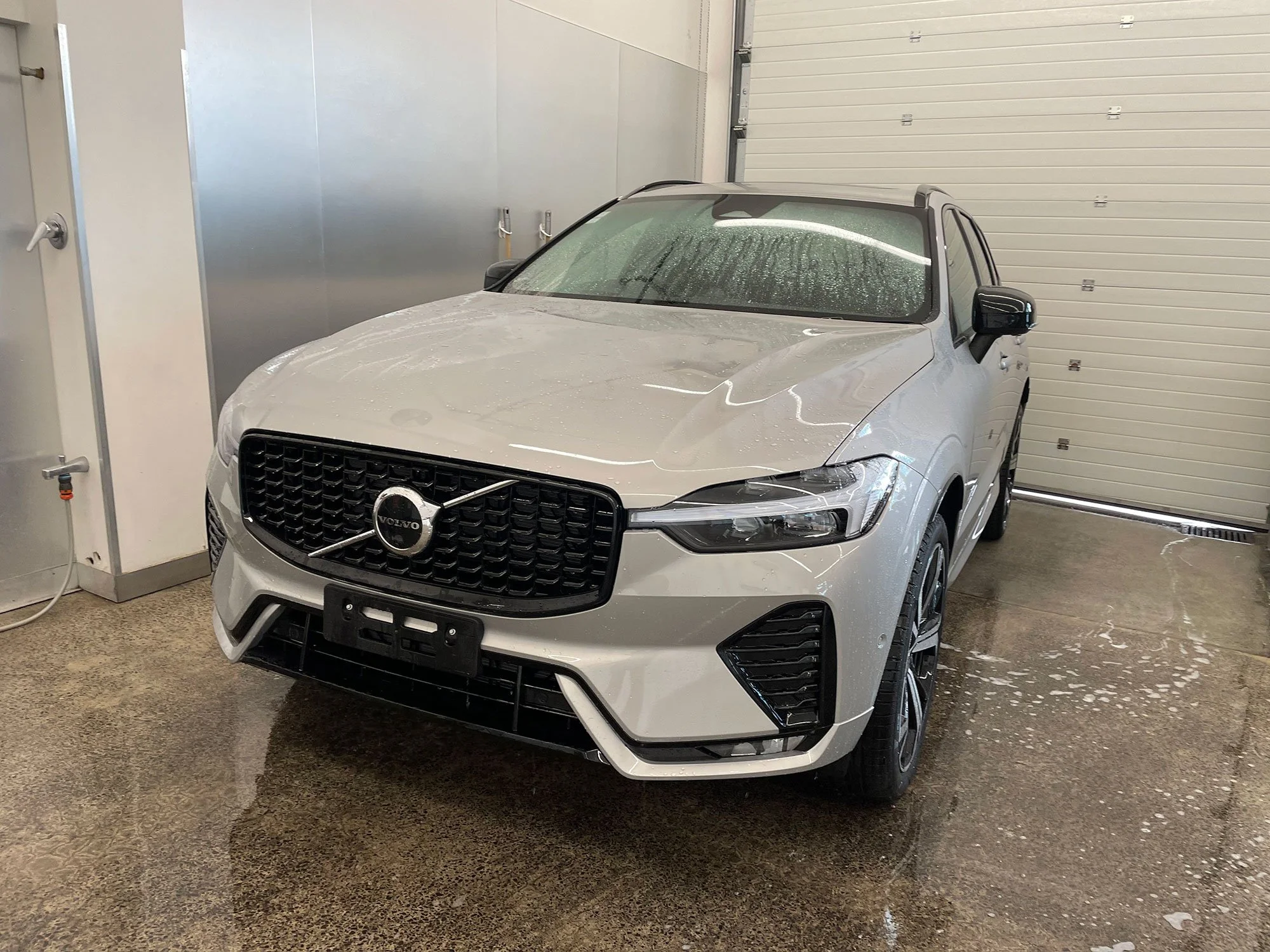 A white Volvo SUV inside a car wash with water droplets on the windshield and front hood, and soap suds on the ground.