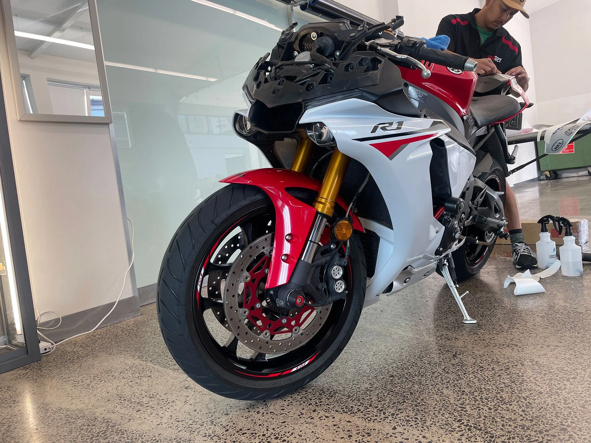 Motorcycle with red, white, and black bodywork and gold front suspension forks in an indoor setting, with a person working on it and cleaning supplies nearby.