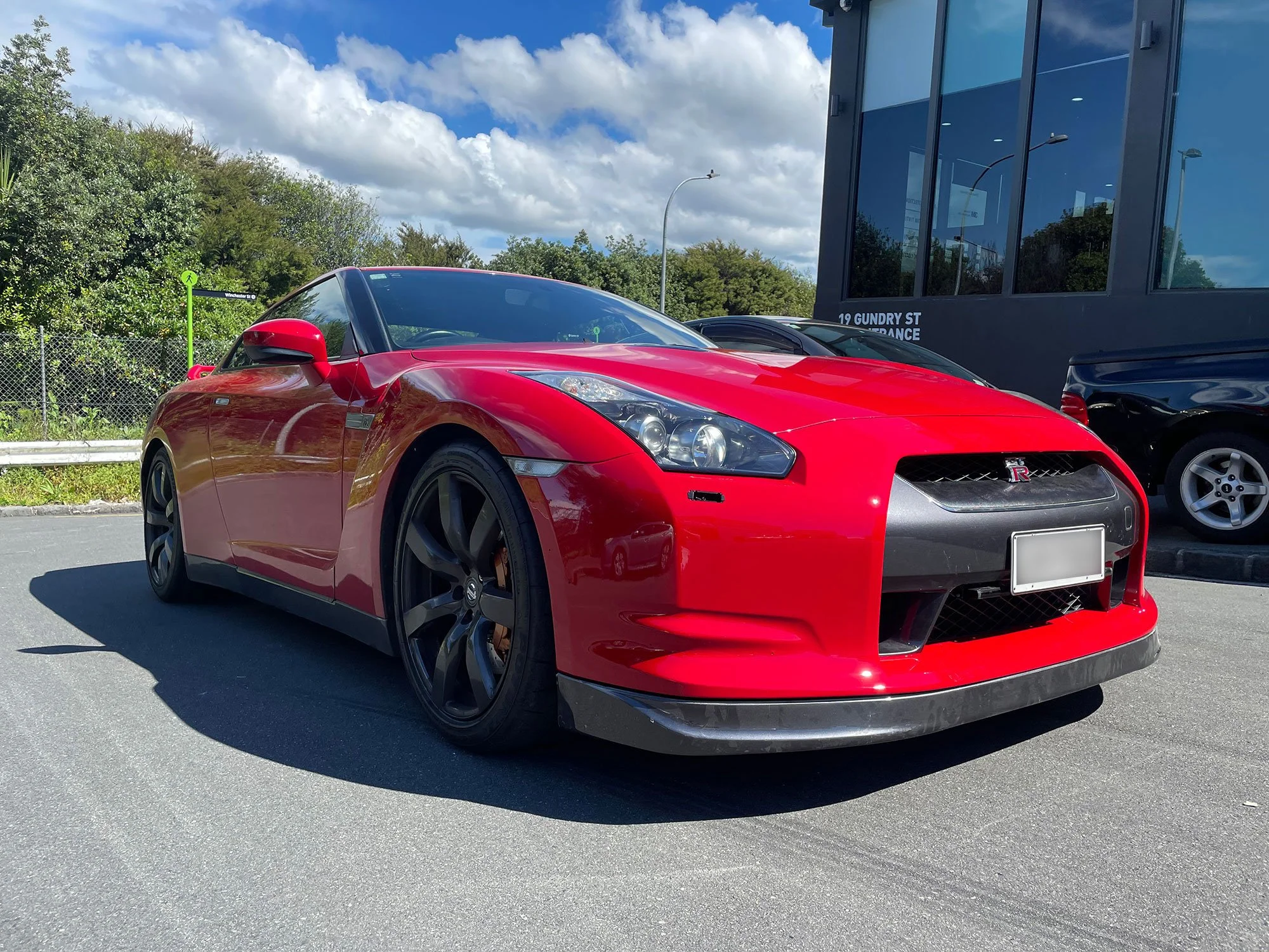 Red Nissan GT-R sports car parked in a lot with a modern building and greenery in the background.