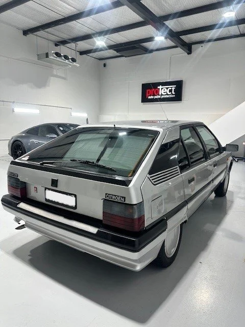 A vintage silver Citroën car parked inside a well-lit garage with a white floor and walls. There is a black and white sign hanging on the wall that reads 'protect auto paint protection'. A modern black car is parked next to the Citroën in the backgro