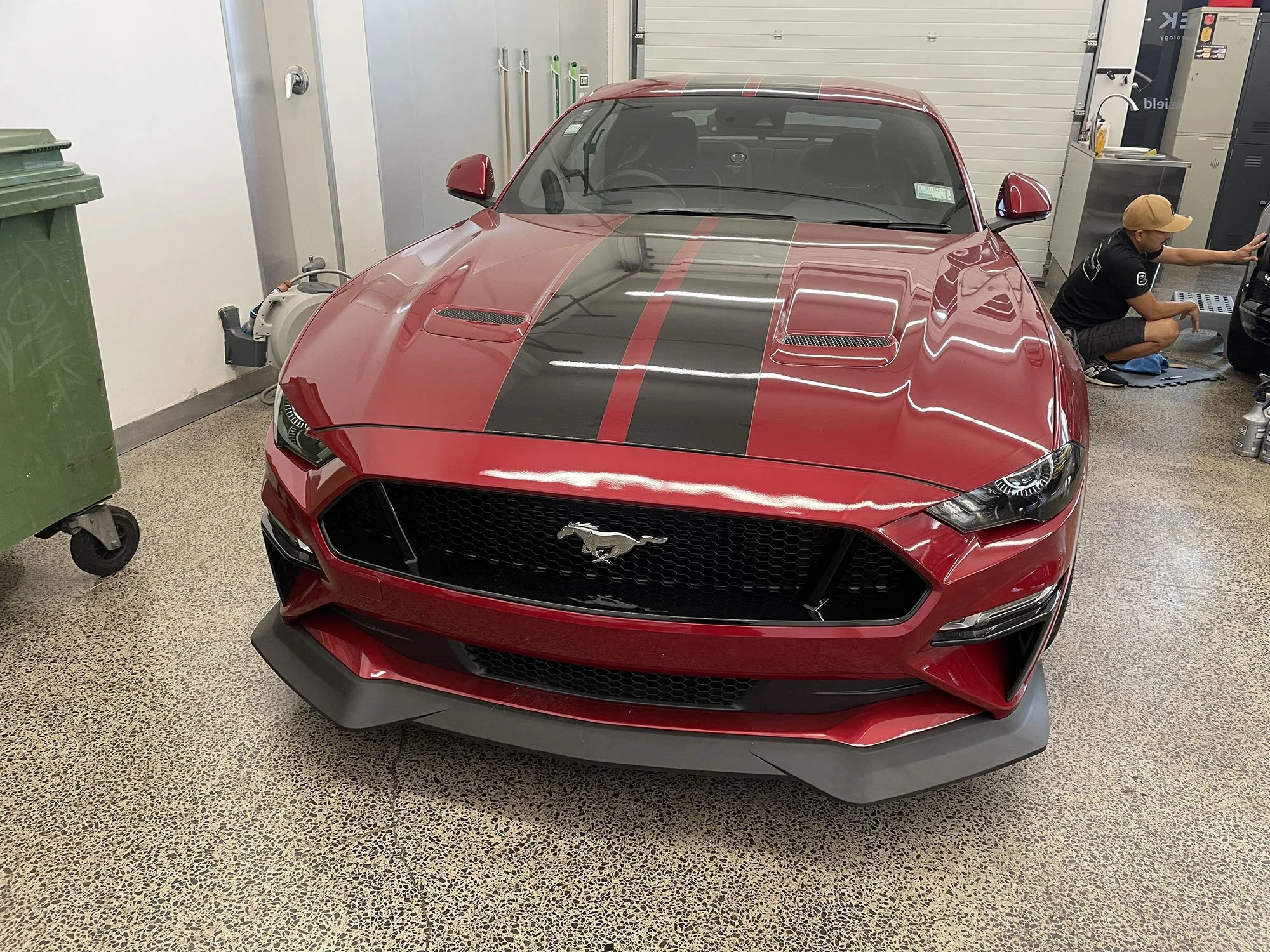 Red Ford Mustang car with black racing stripe in a garage, with a person working on a computer nearby.