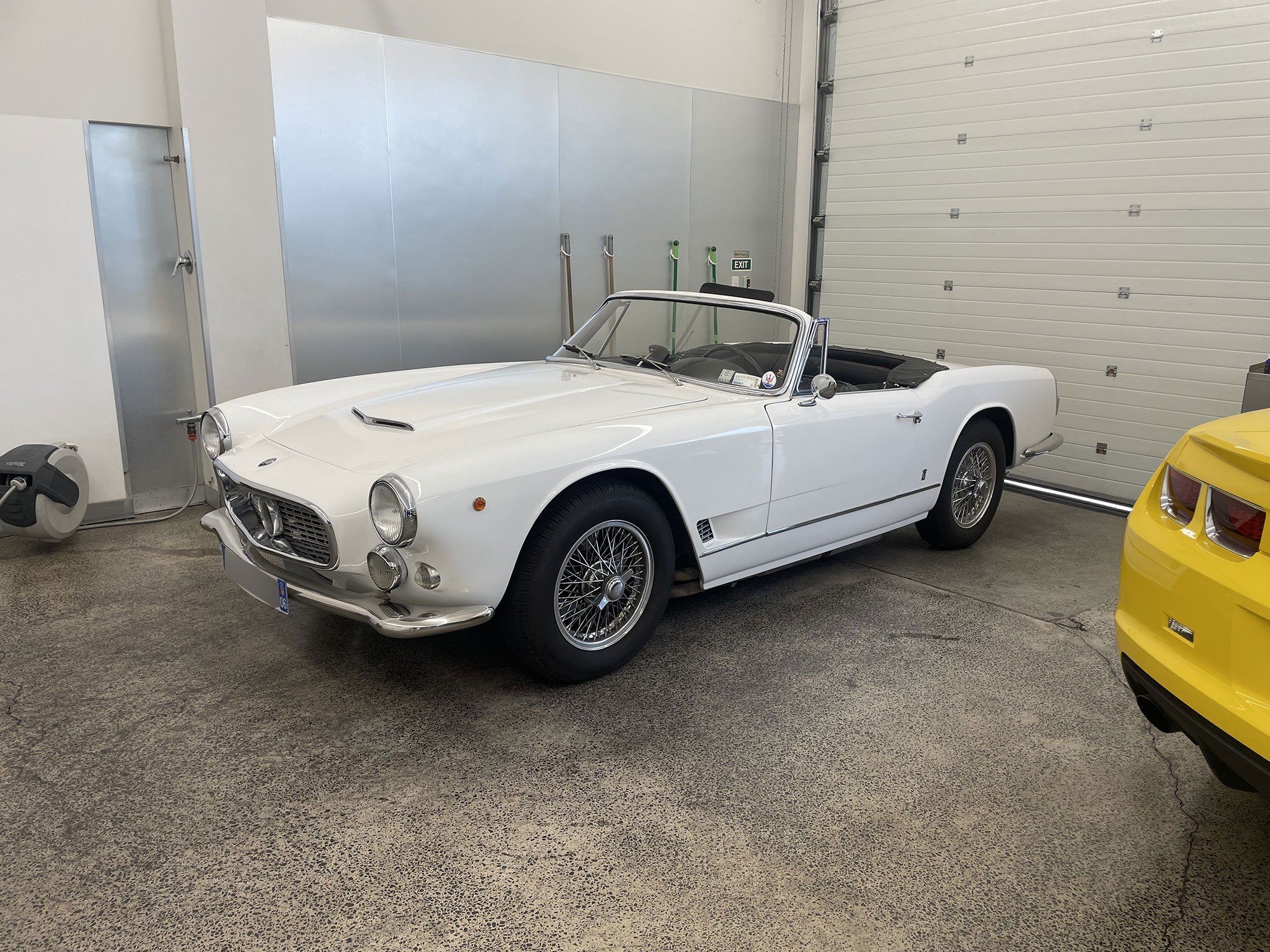 Classic white convertible sports car inside a garage, parked on a concrete floor next to part of a yellow vehicle.