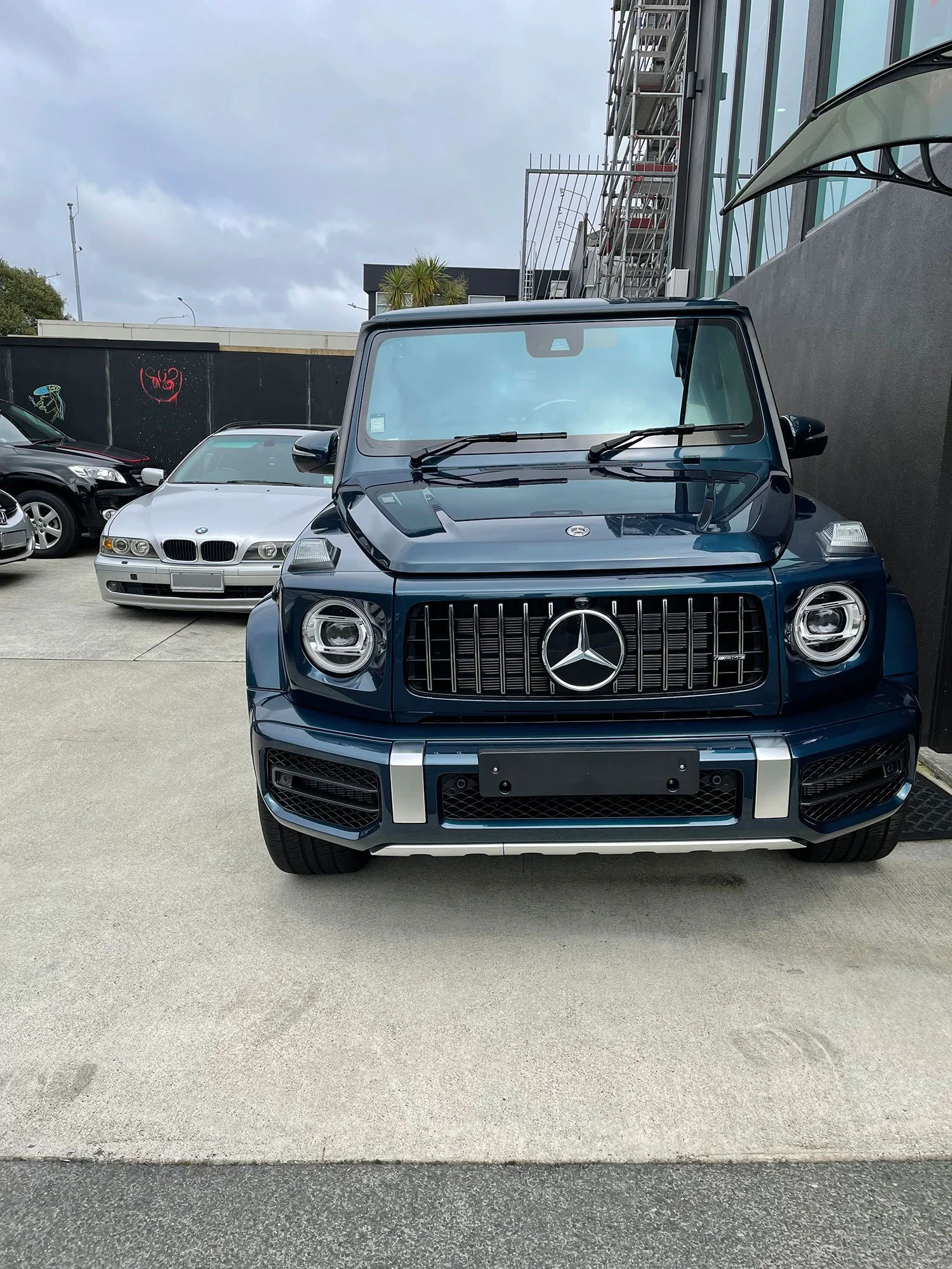 Front view of a dark blue Mercedes-Benz G-Class SUV parked in a lot, with a white BMW and other cars nearby, against a background of buildings and a cloudy sky.