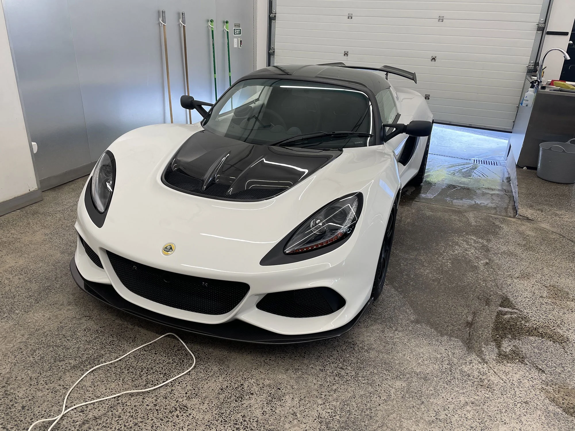 White and black Lotus sports car parked inside a garage with a closed garage door and various tools hanging on the wall.