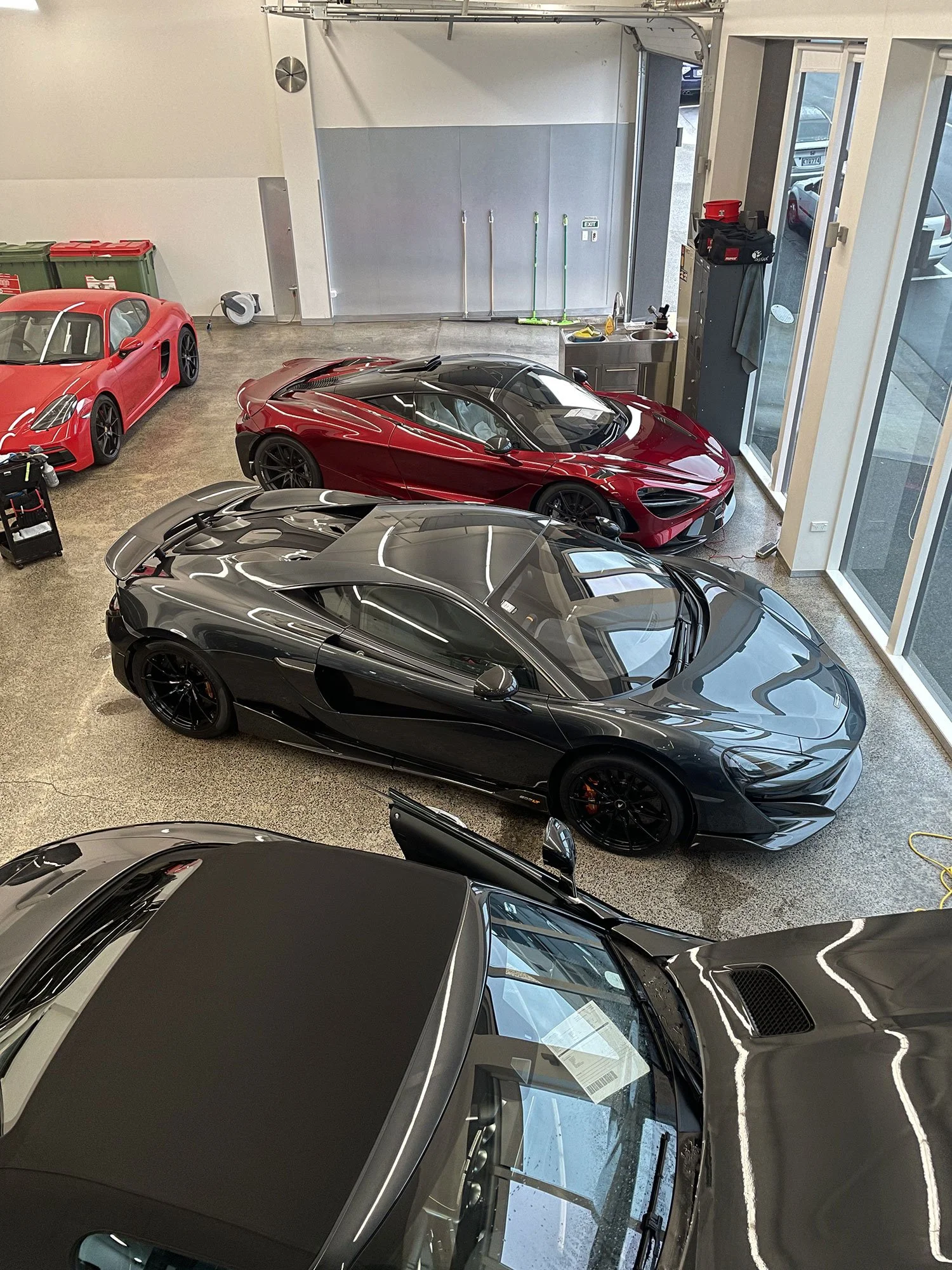 A showroom with luxury sports cars, including a black McLaren, a red McLaren, and a red Ferrari, parked inside near large glass windows.