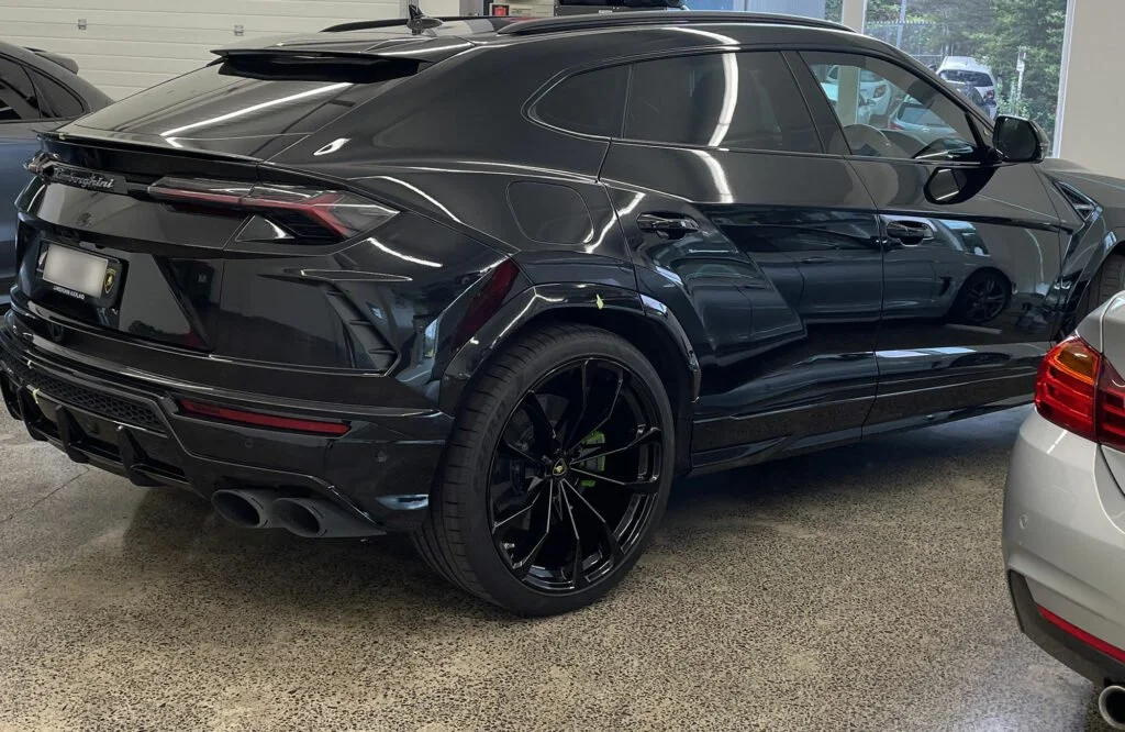 Black Lamborghini Urus parked indoors with other cars visible nearby.