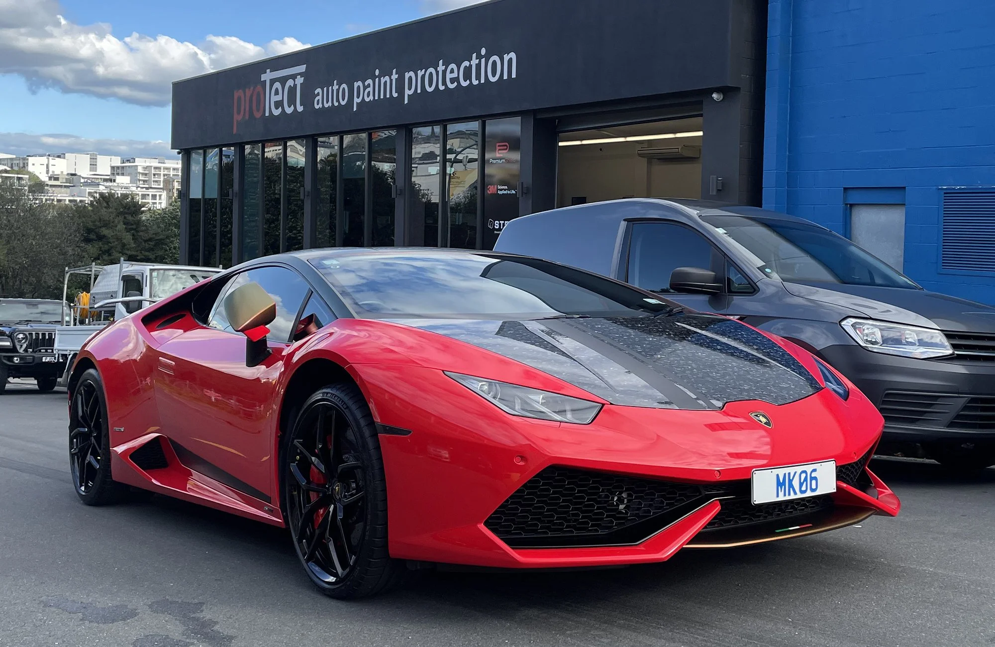 A red Lamborghini Huracan with black wheels parked outside an auto paint protection shop, with buildings and a cloudy sky in the background.