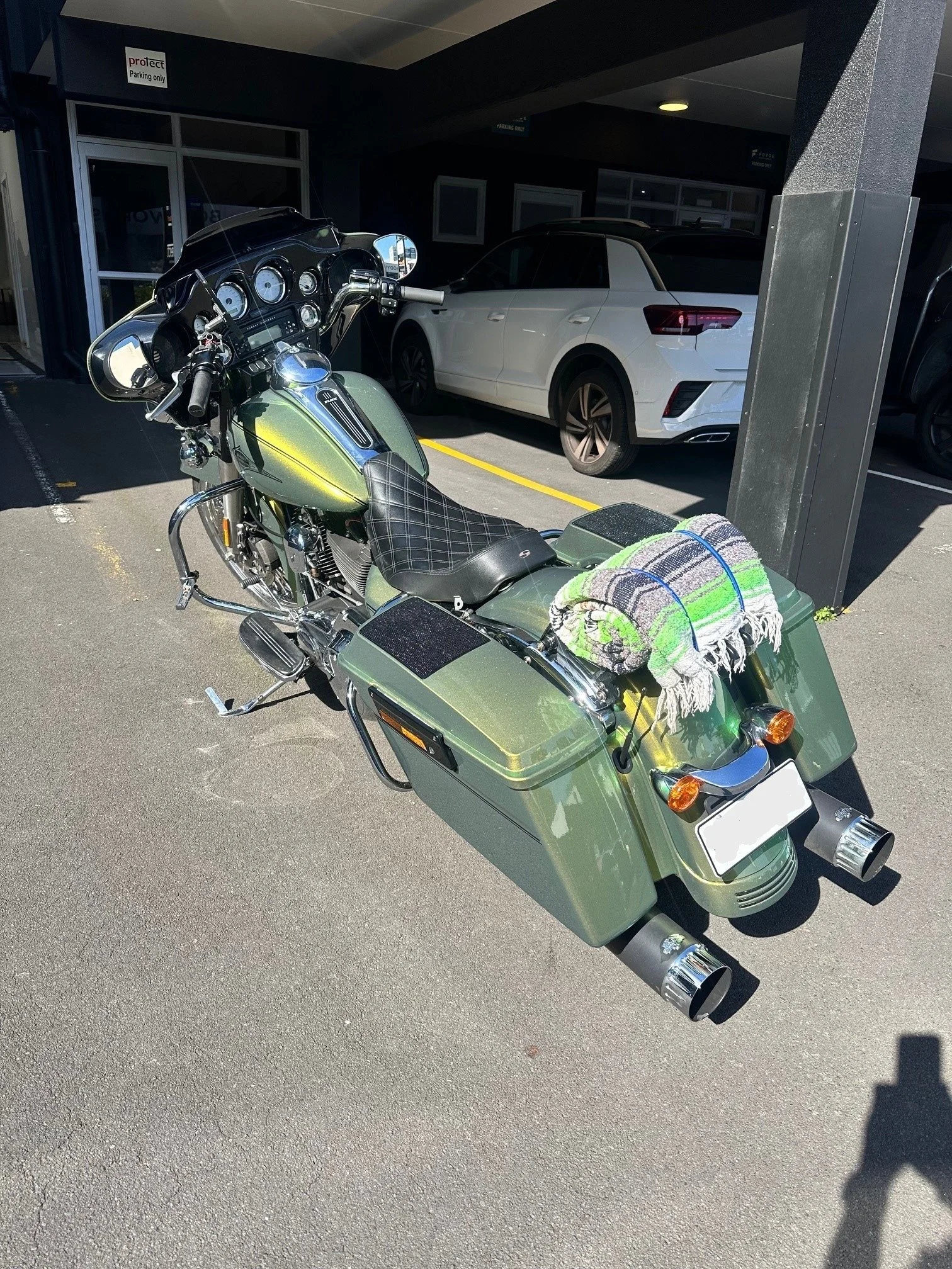 A green touring motorcycle with saddlebags parked in a parking lot, with a towel draped over the back, and a white car behind it.