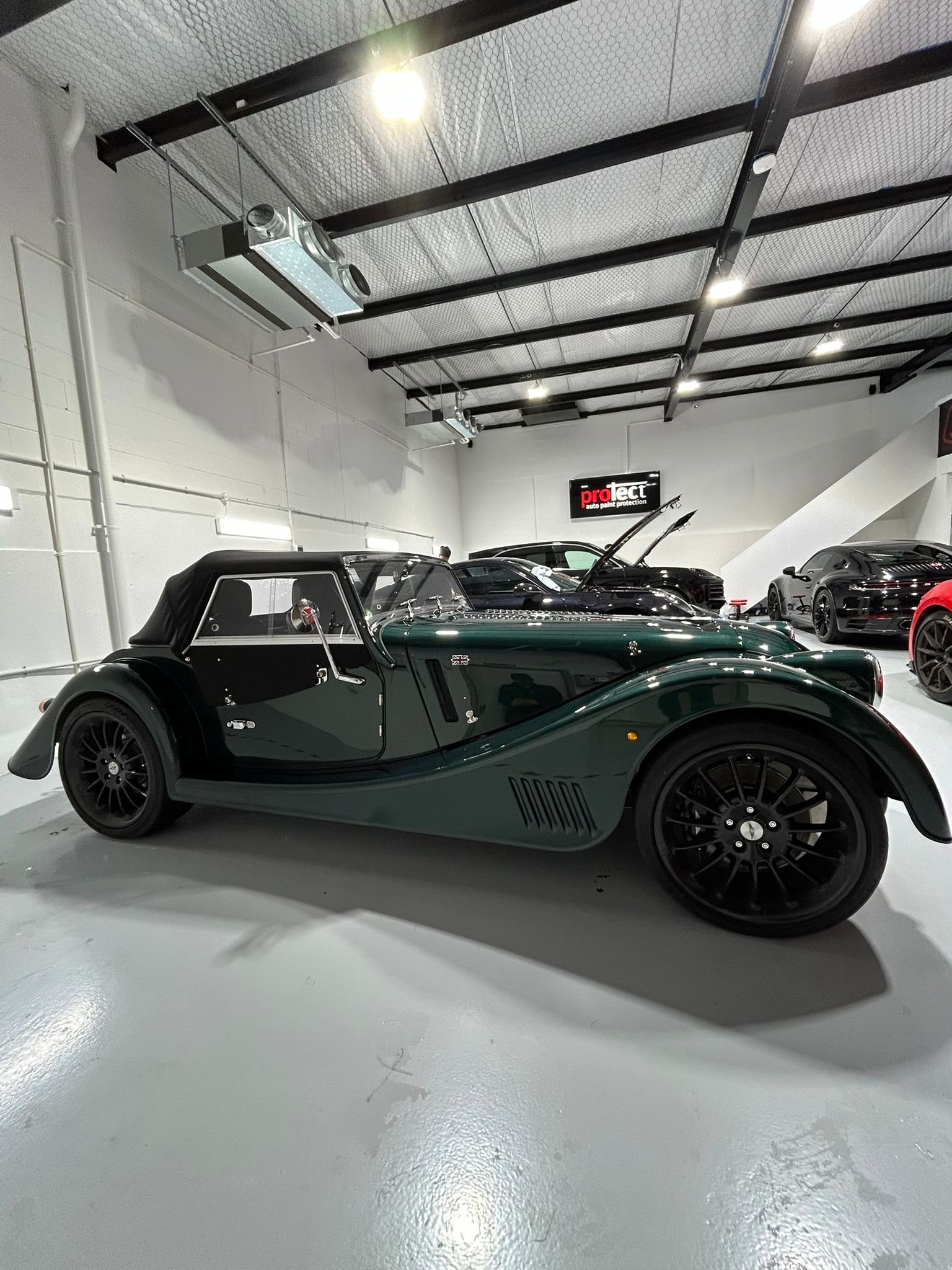 A vintage dark green convertible sports car is displayed in an indoor car showroom, surrounded by other modern cars.