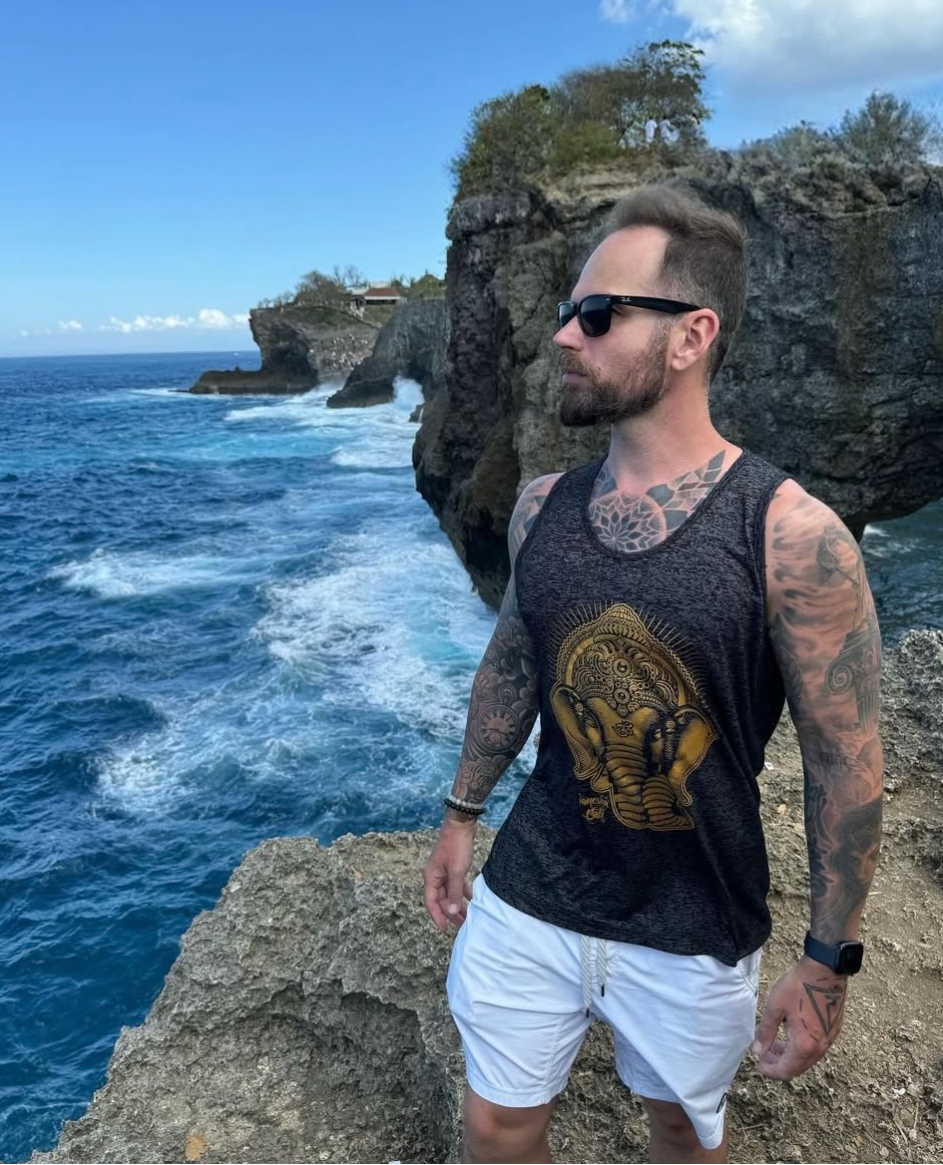 A man with tattoos wearing sunglasses, a black tank top with a golden elephant design, and white shorts standing on rocky cliffs overlooking the ocean, with waves crashing below and clear blue sky.