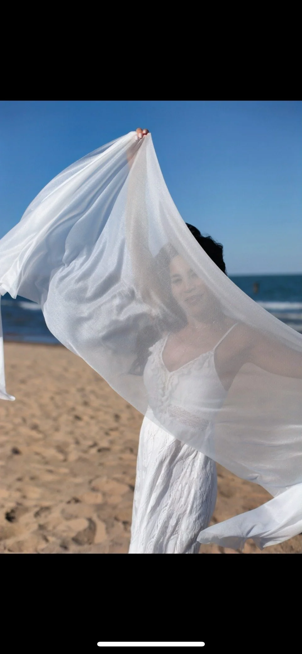 A woman wearing a white dress peaking behind a veil.