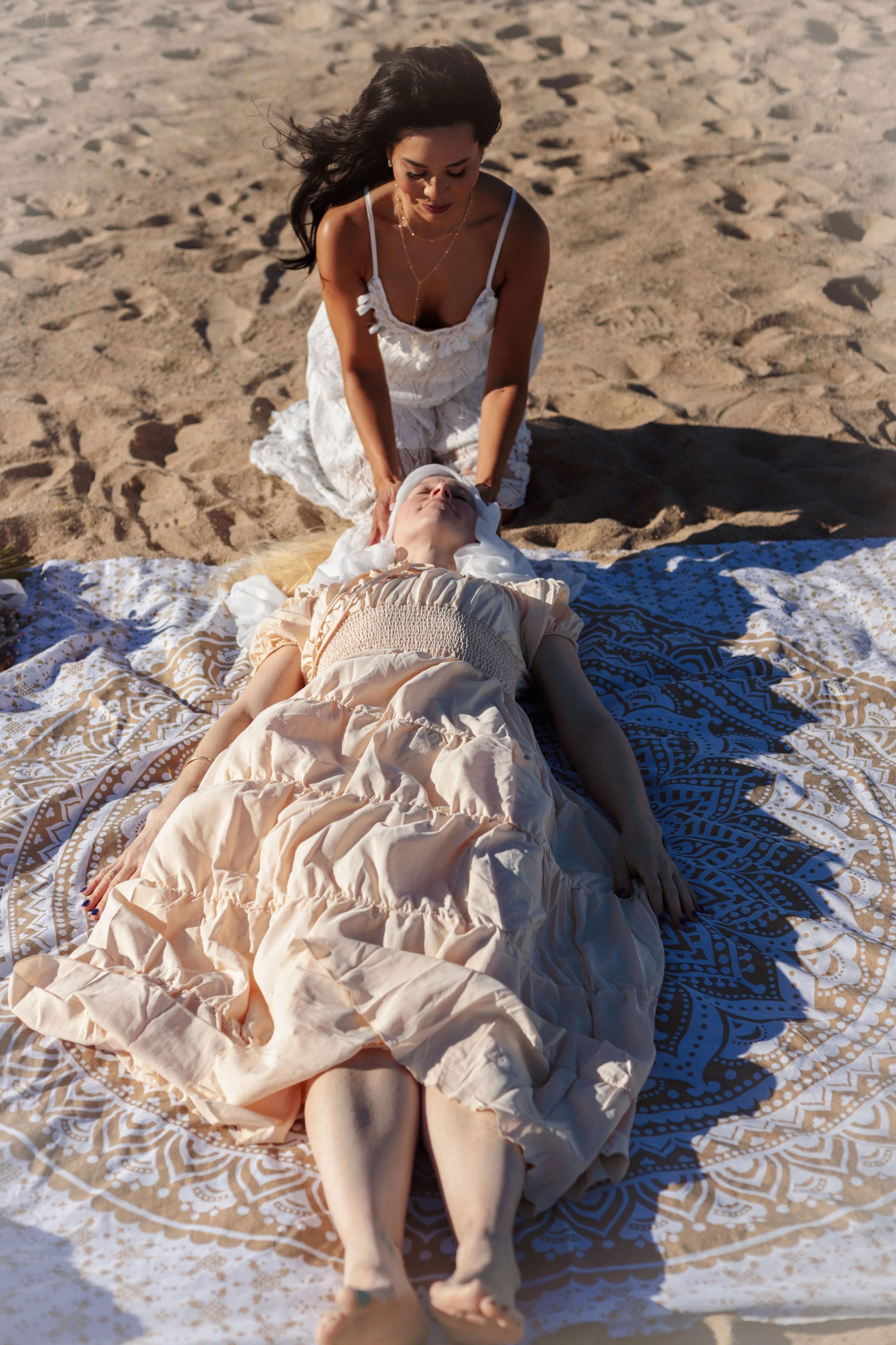 A woman is giving a spiritual or ceremonial blessing or prayer to another woman lying on a patterned blanket on the sand at the beach.