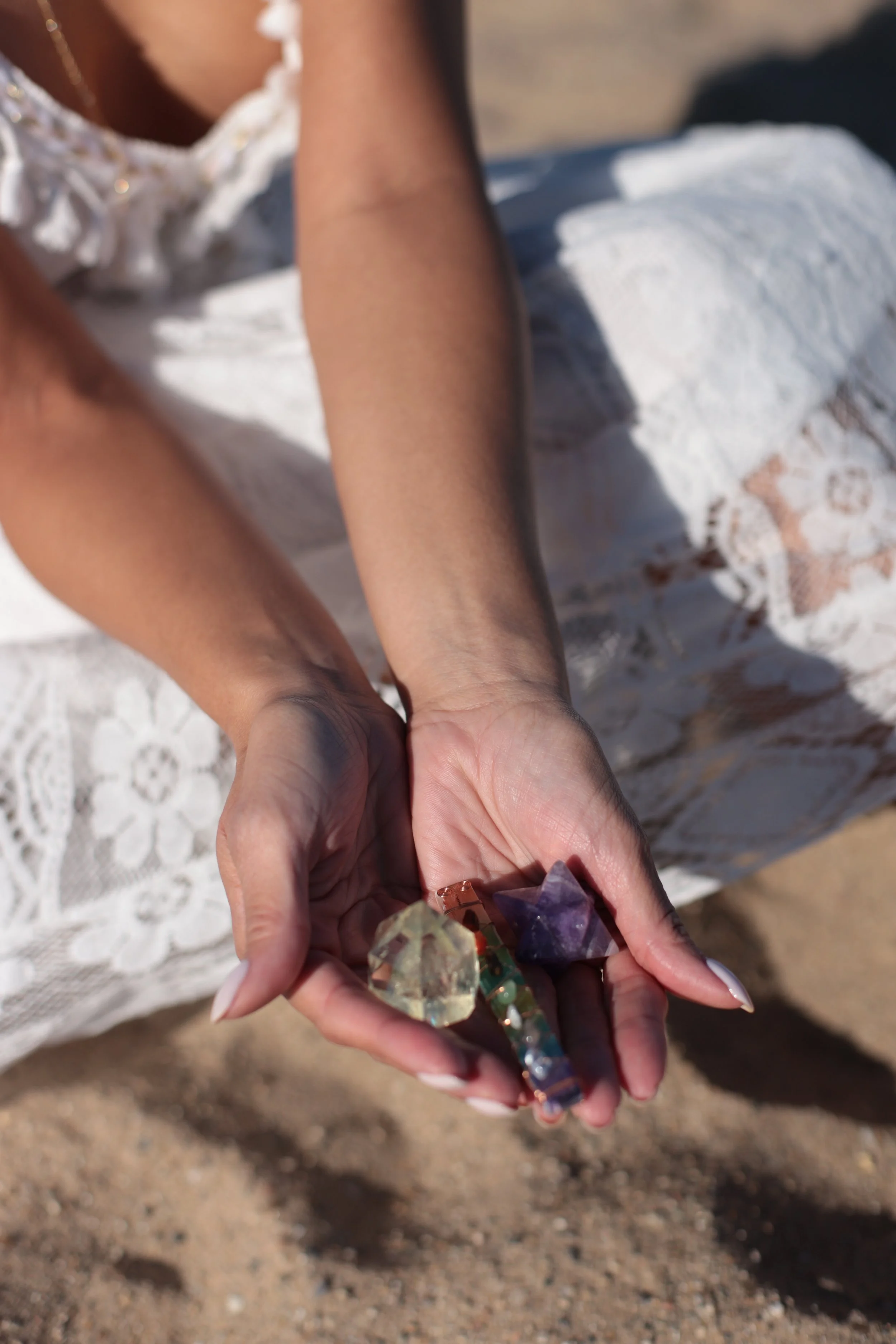 Person holding a collection of colorful crystals, wearing a white lace dress, with sandy ground beneath.