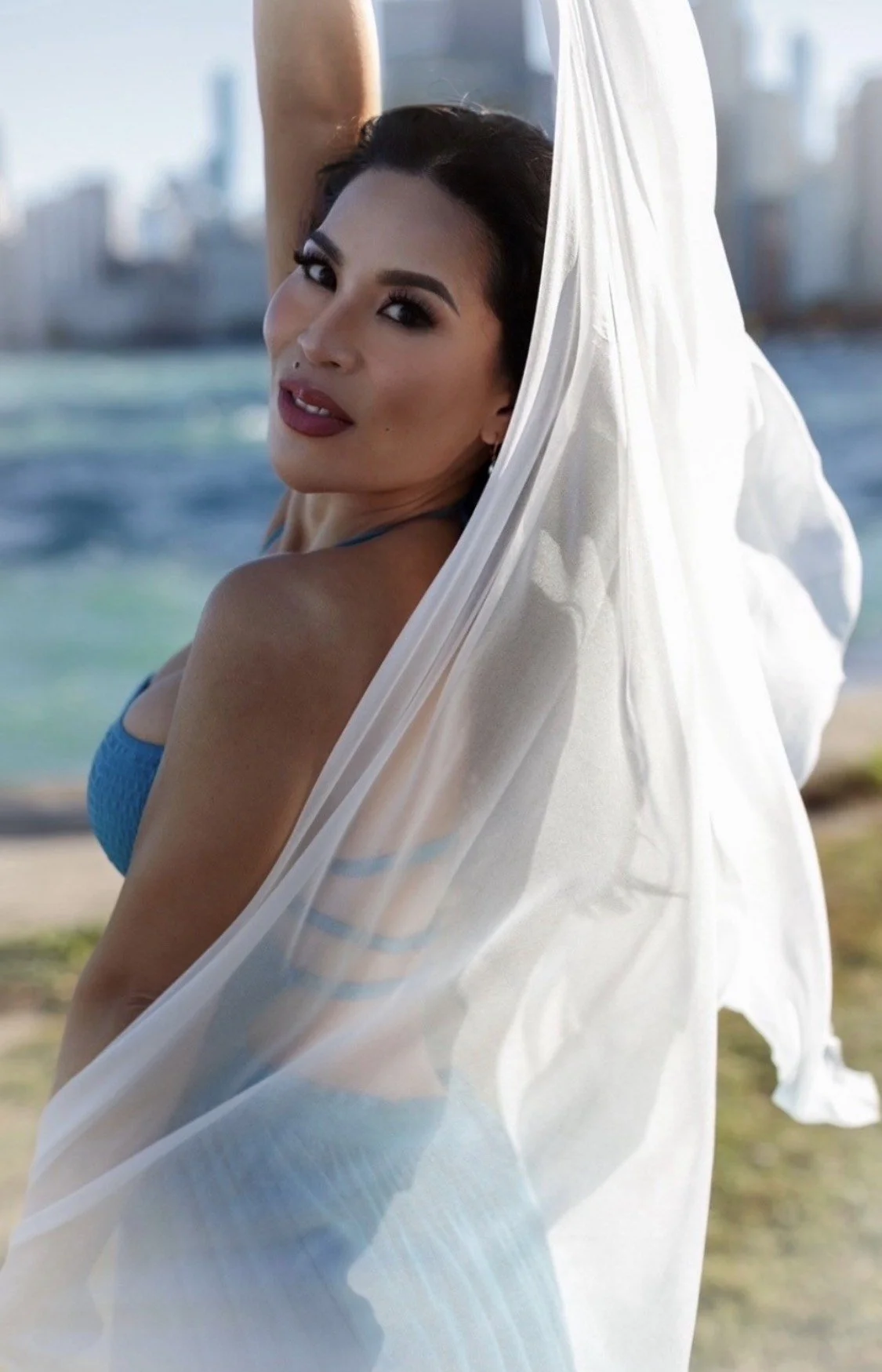 A woman with dark hair and makeup, wearing a blue swimsuit, stands outdoors near water with a city skyline in the background. She holds a sheer white fabric draped around her.