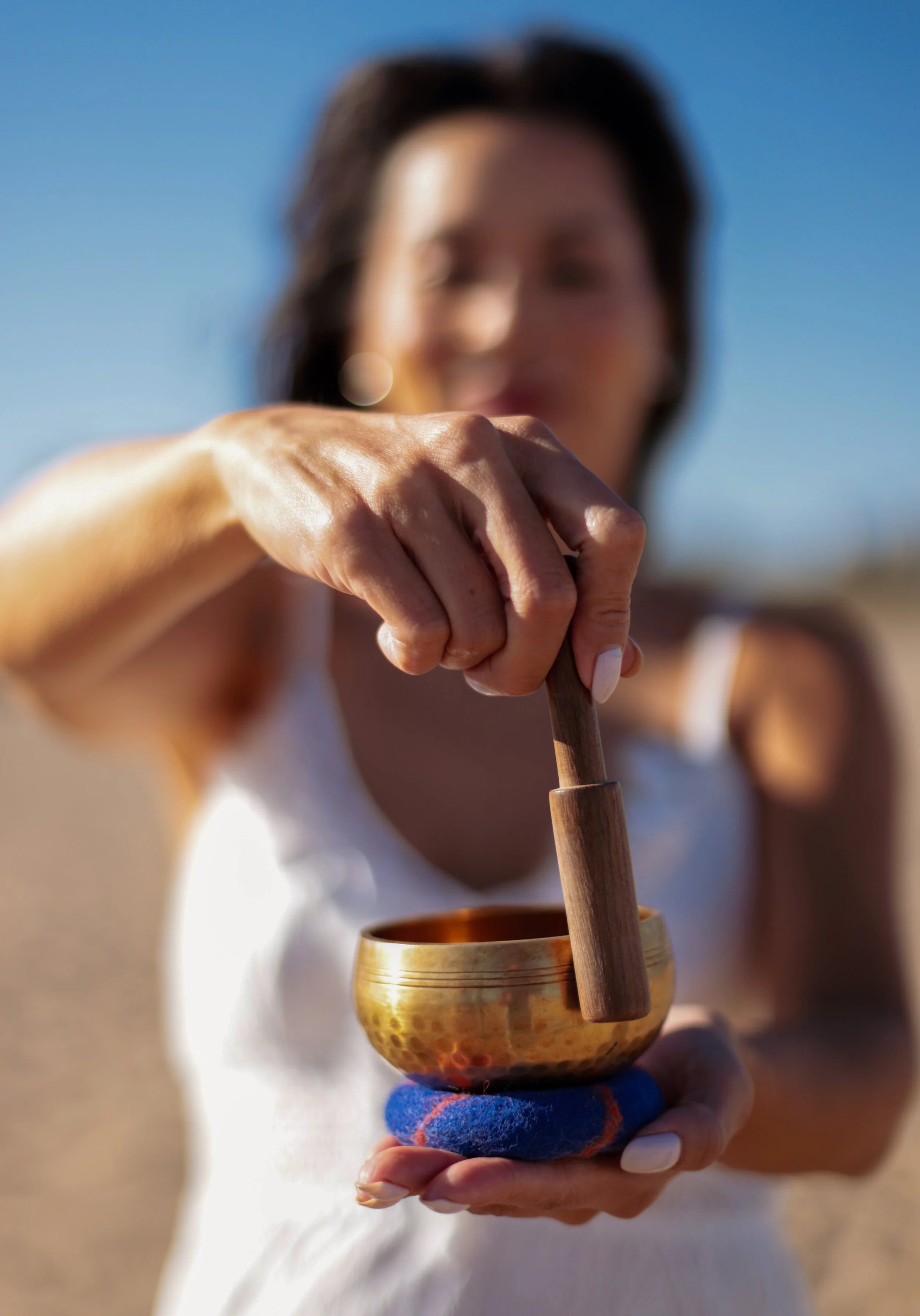 A woman is holding a brass singing bowl and a wooden mallet, with a blue felt pad between her hand and the bowl, outdoors under a clear blue sky.