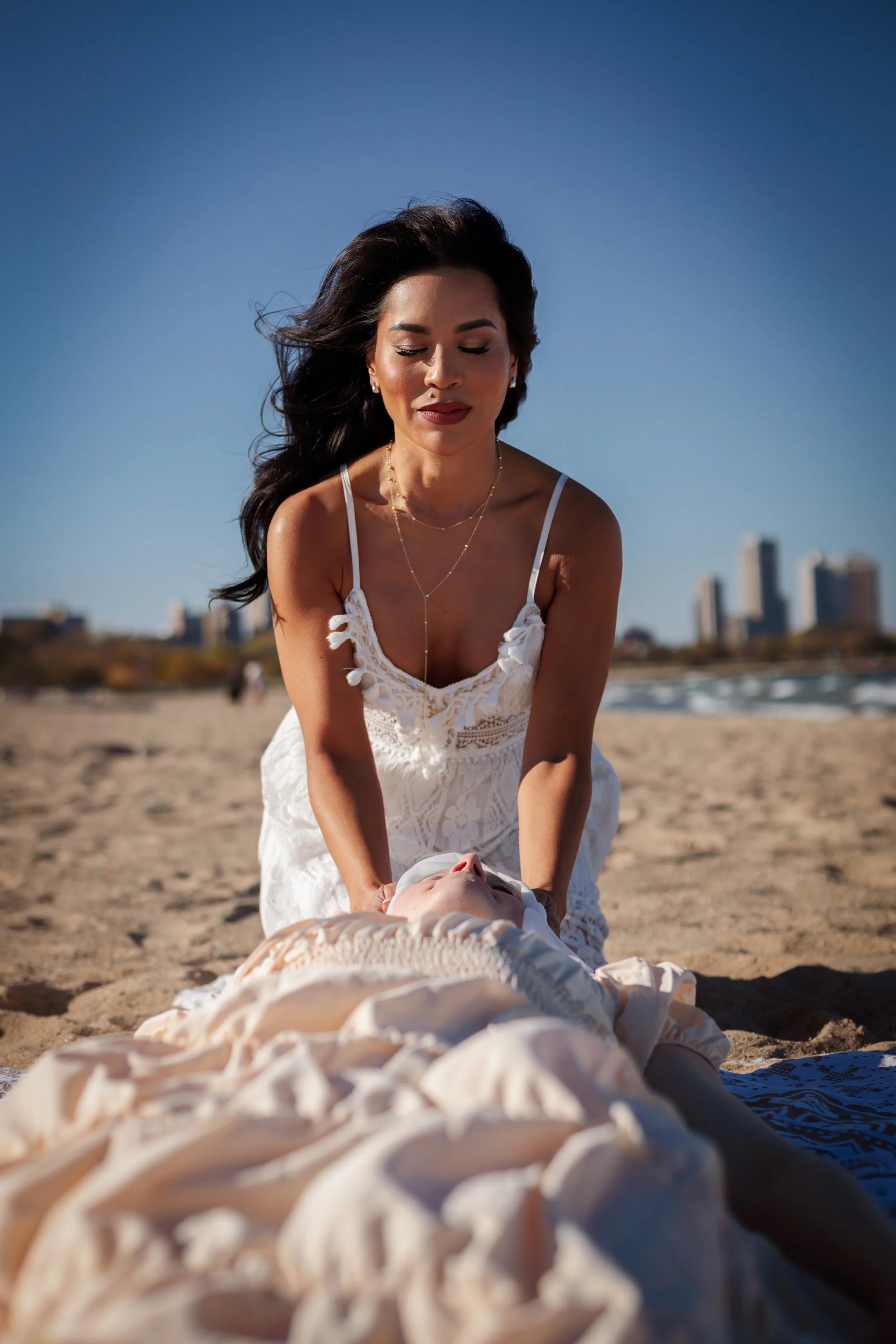 A woman in a white dress giving a massage on the beach with city buildings in the background.