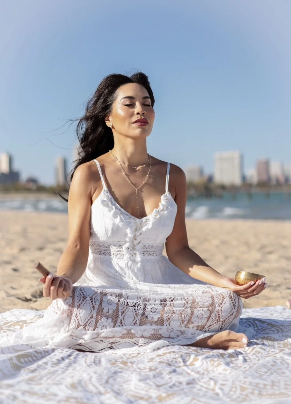 A woman practicing meditation on a beach, sitting cross-legged on a blanket, holding a singing bowl and a meditation stick.