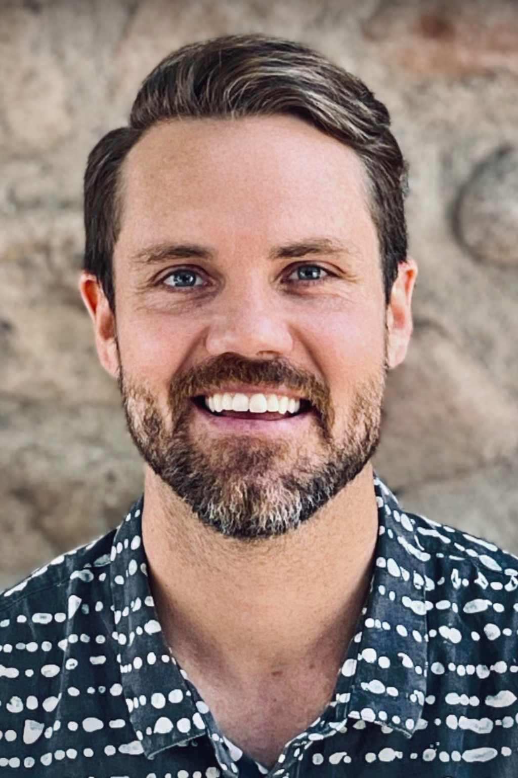 A smiling man with short brown hair and a beard, wearing a patterned black and white shirt, standing in front of a stone wall.